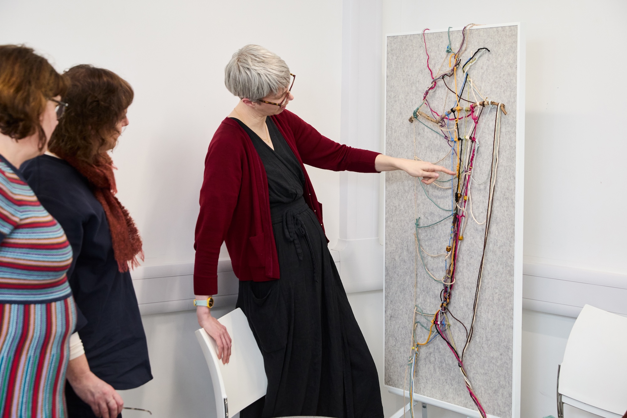A woman with short grey hair points to an abstract wall sculpture, while two other women stand beside her in a white gallery space.