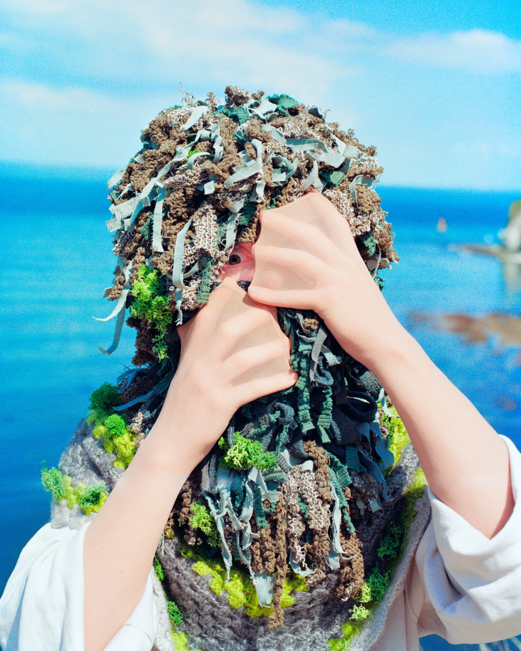 A person covers their face with hands, wearing a textured green and brown mask made of fabric and natural materials. The background features a blue sea under a clear sky, with rocky shorelines visible.