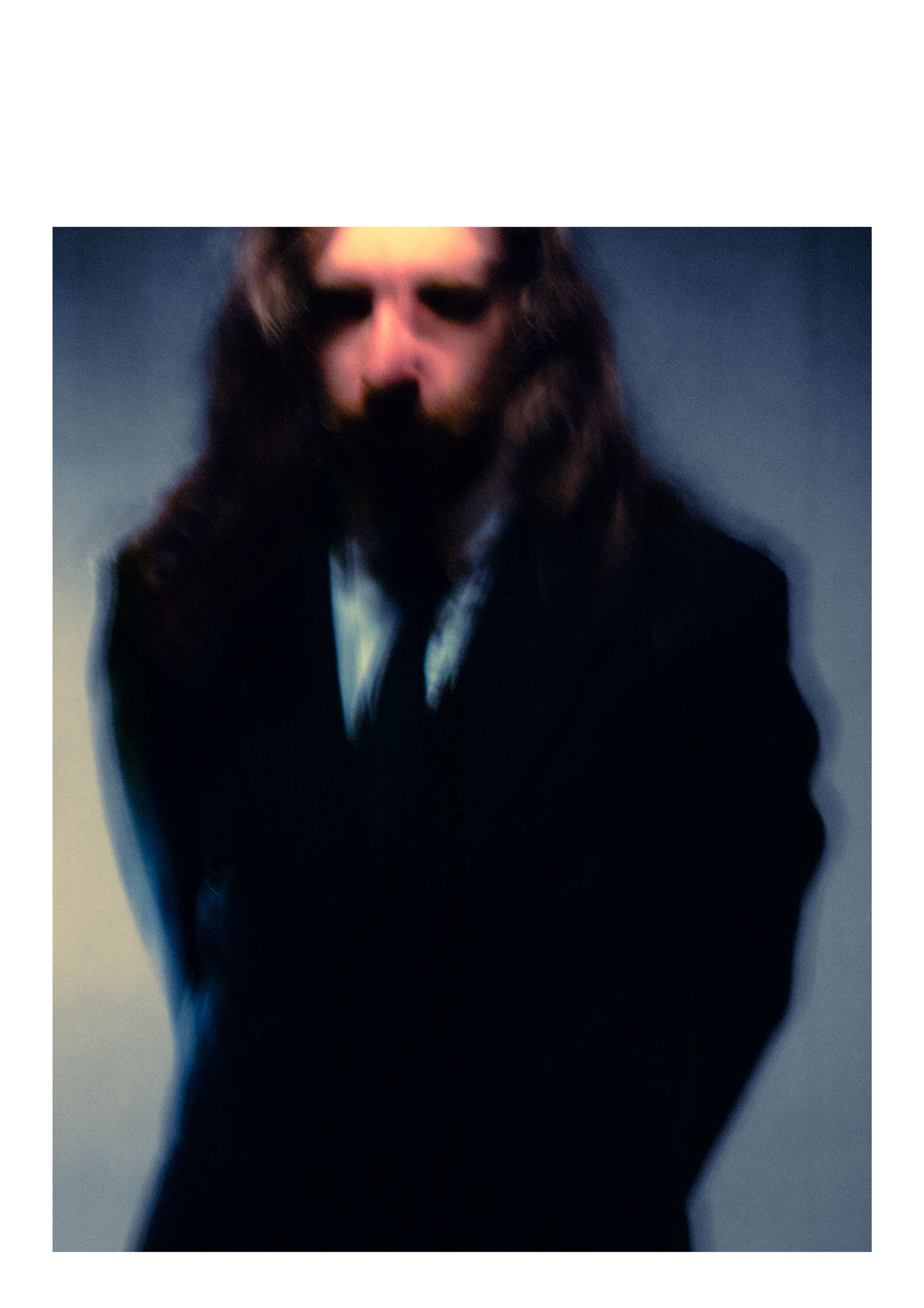 A person with long hair, dressed in a black suit, stands with their hands behind their back. The image is out of focus, creating a blurred effect, and the background is a gradient from light to dark.