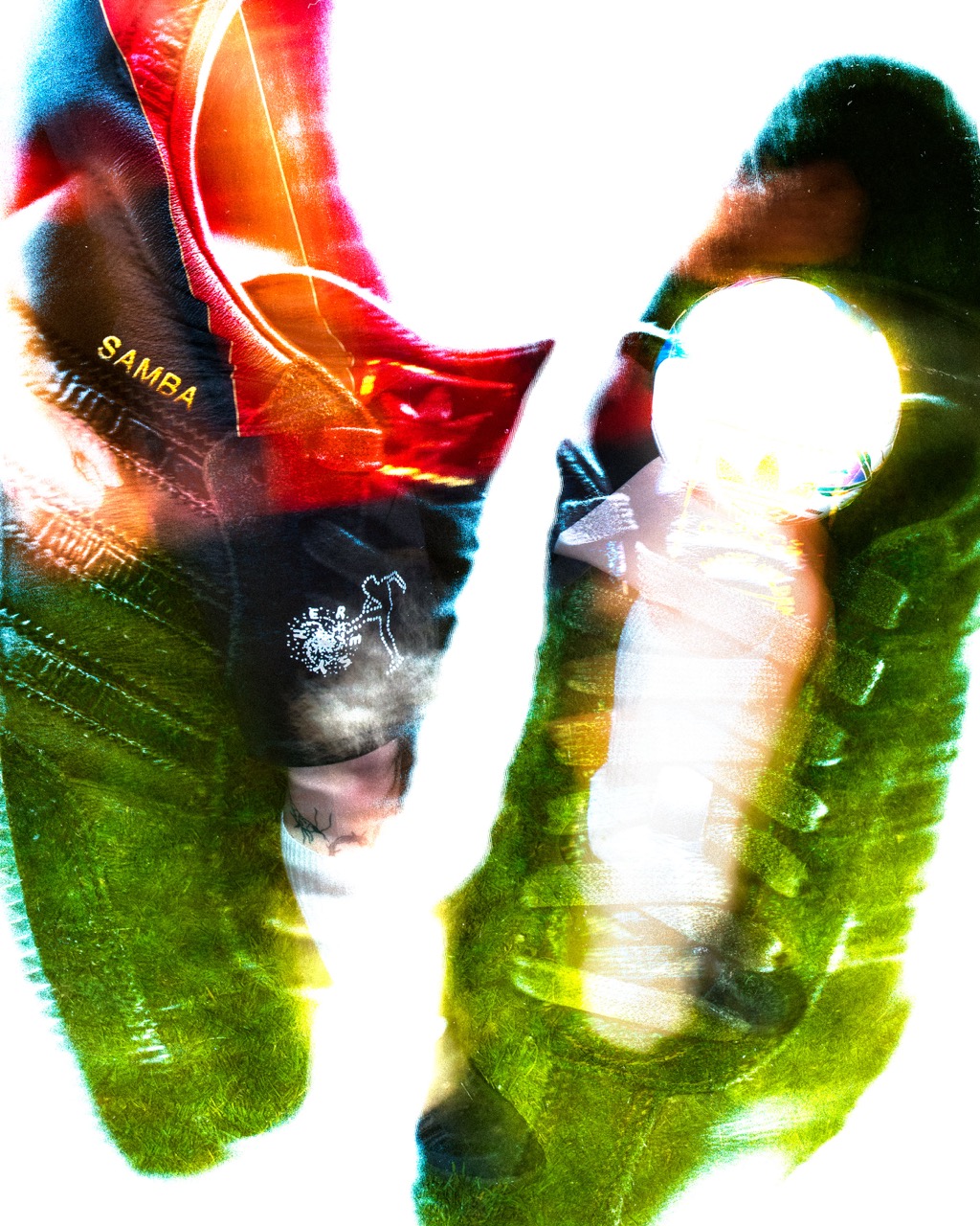 A pair of football boots and a football, photographed against a bright background. The boots are partially obscured, one is red with the word "SAMBA" visible, while the other is black with a light reflection.