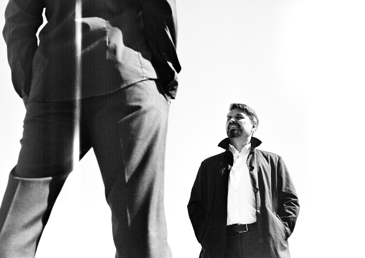A man in a dark coat looks up at another man standing in front of him. The foreground features the legs of the standing man, while the background is white, highlighting the contrasting figures.