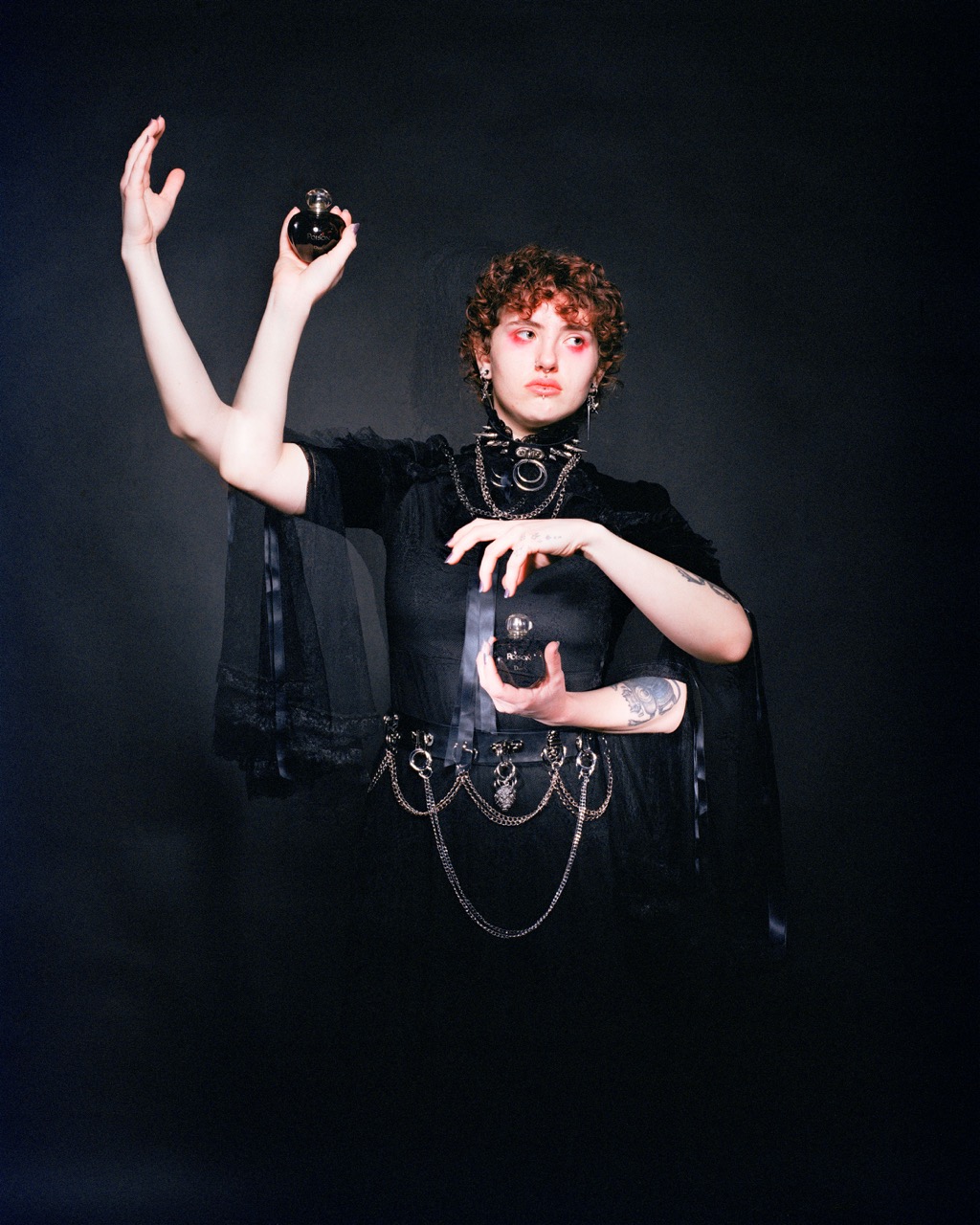 A person with curly hair wearing a black outfit and chains stands against a dark background, holding two small bottles. Their raised left arm holds a bottle, while the right hand grips another one closer to their body.
