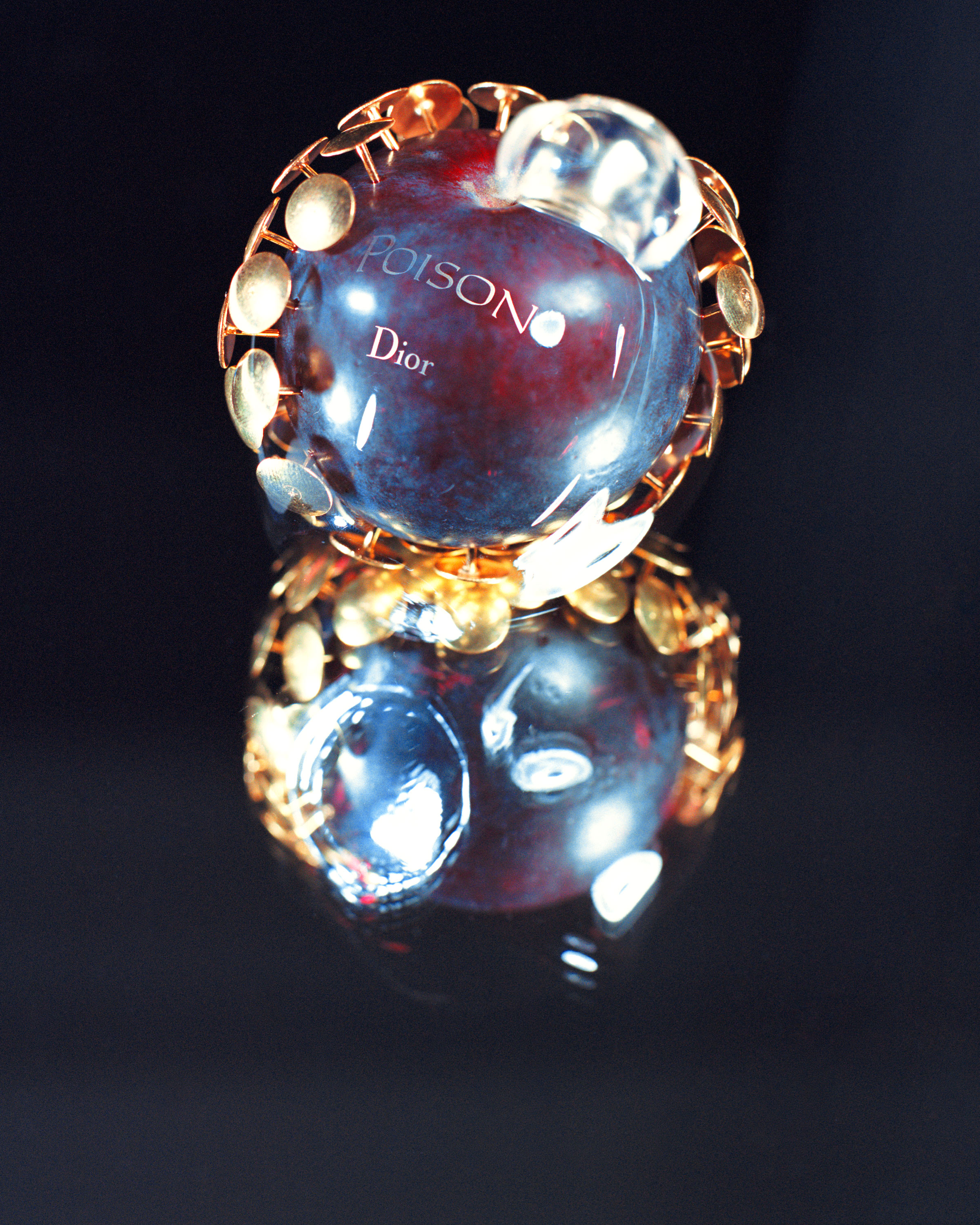A round glass perfume bottle with a gold and transparent cap sits on a reflective surface. The bottle is predominantly dark red with the word "Poison" and "Dior" visible on the front.