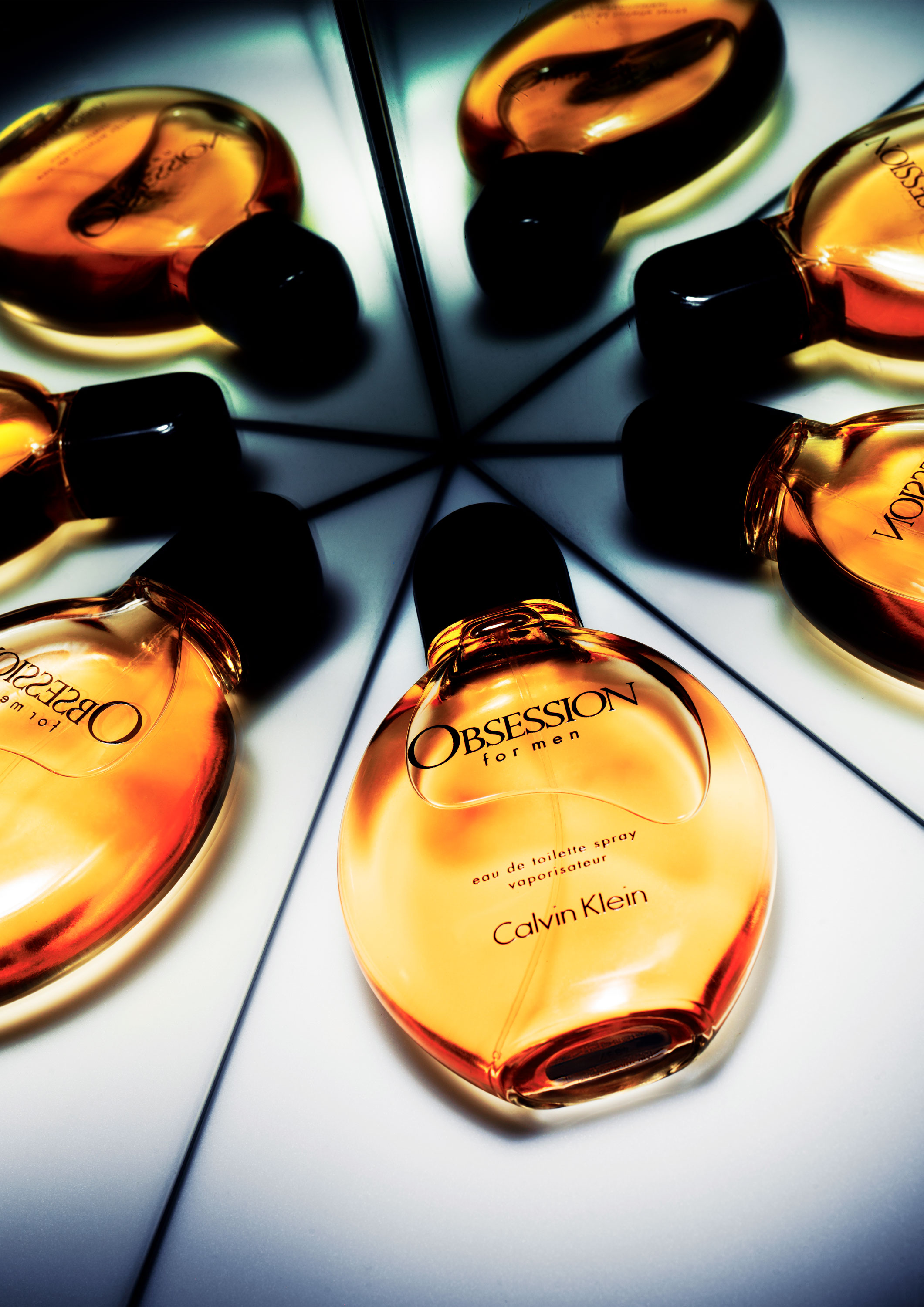 A bottle of Calvin Klein's 'Obsession for Men' eau de toilette placed on a reflective surface, surrounded by multiple reflections of the bottle. The glass bottle is a dark amber colour with a black cap and label.