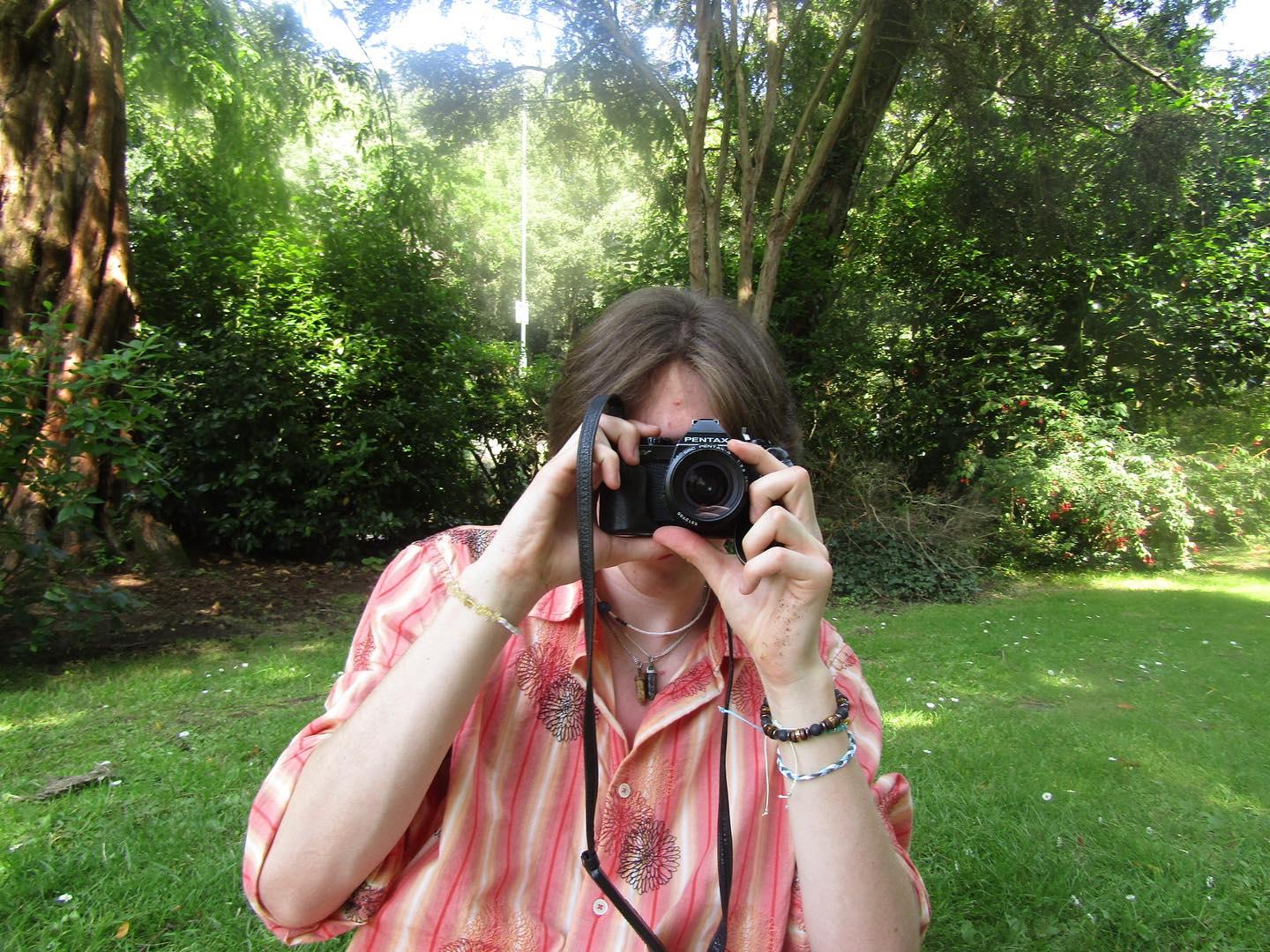 A person in a pink striped shirt holds a camera to her face in a leafy garden, with grass and trees in the background.