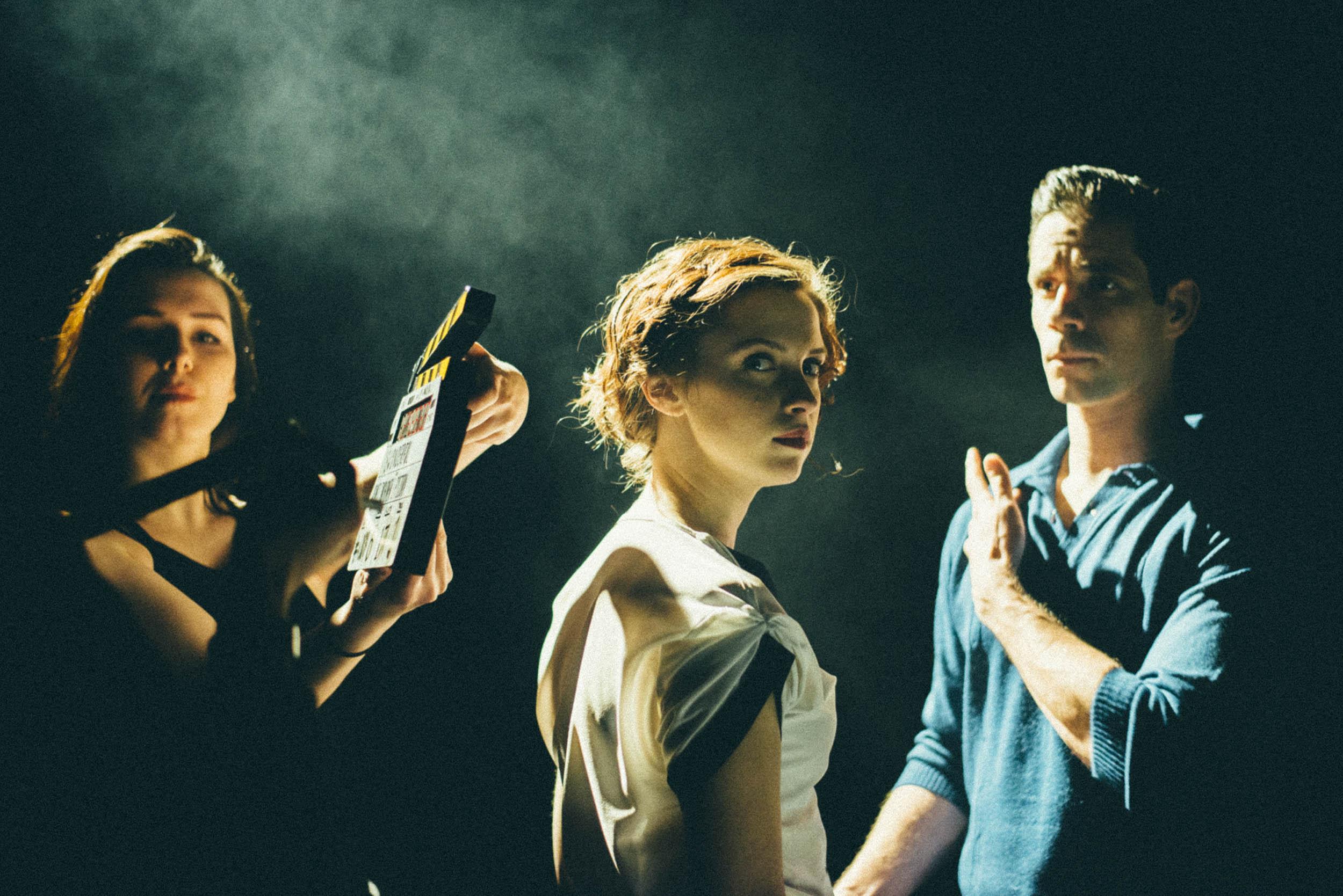 Two actors lit by a spotlight. A person standing to the left holds a clapper board out in front of them.