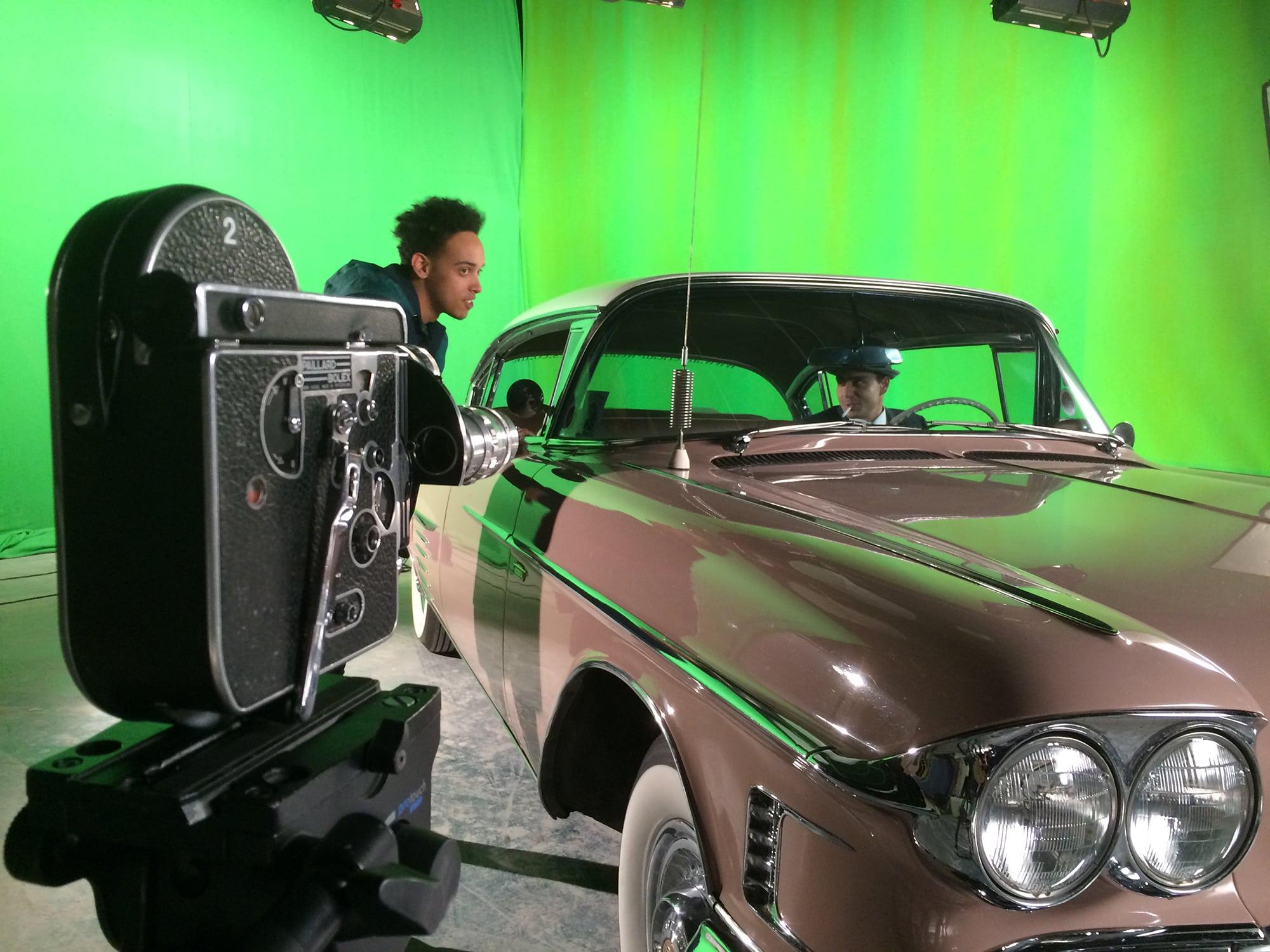 A film camera directed at a vintage car with two people, one outside the car, the other inside. A green screen is on the background.