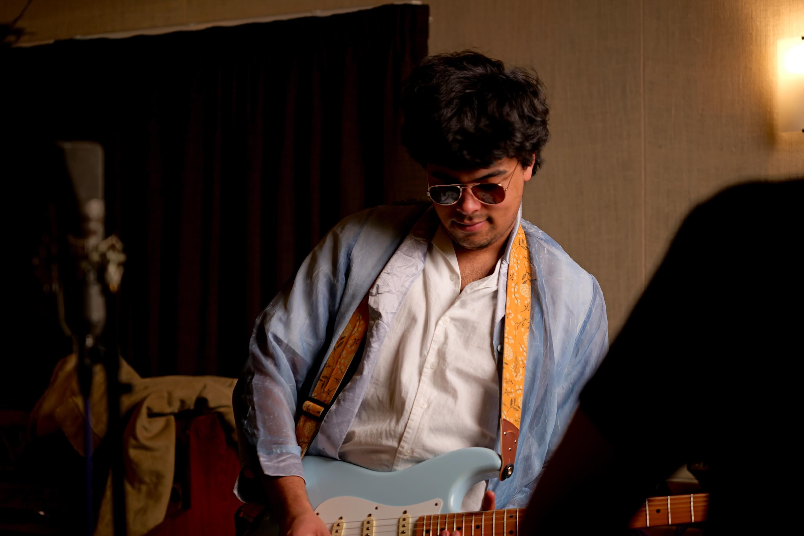 A young person dressed in blue and white with tinted round glasses plays a light blue electric guitar.