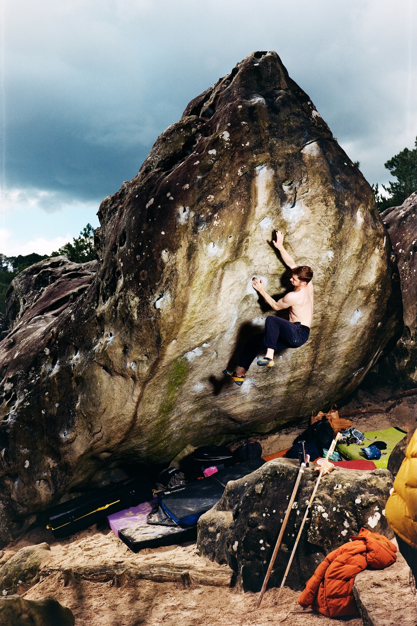 Rock climber scaling large boulder outdoors with crash mats and climbing gear scattered below on rocky ground.