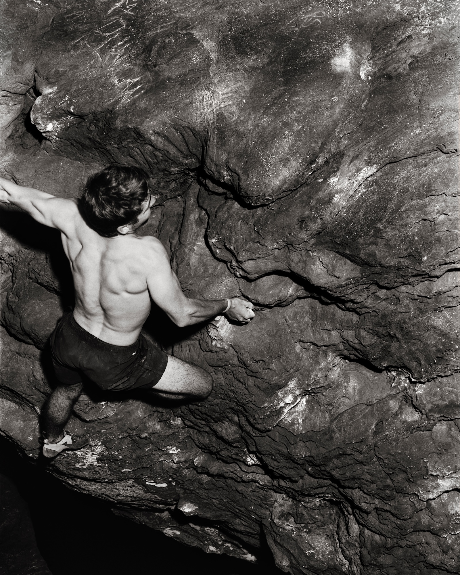Shirtless man in dark shorts climbing on grey rock face with green moss patches and visible cracks in the stone surface.