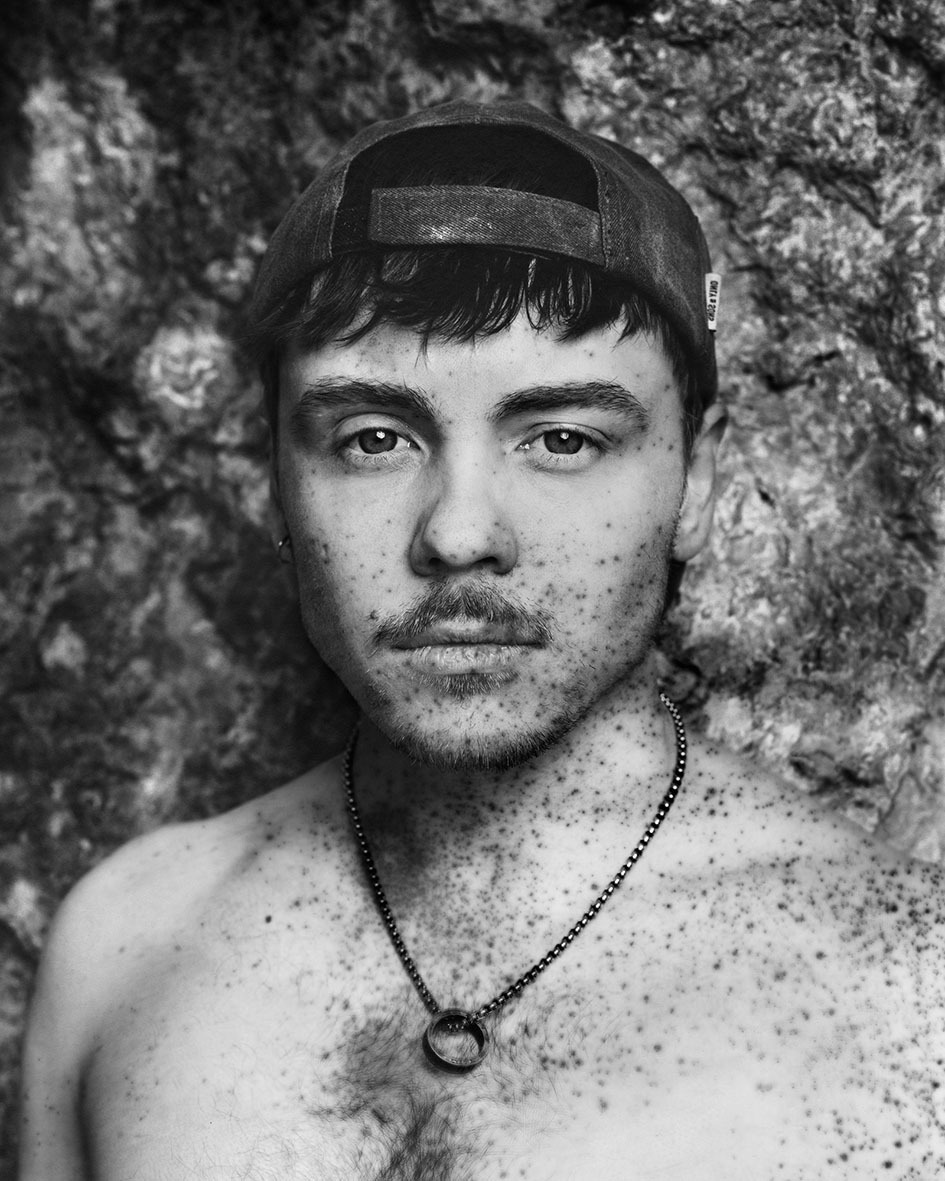 Black and white portrait of shirtless man wearing backwards cap and necklace against textured rock wall background.