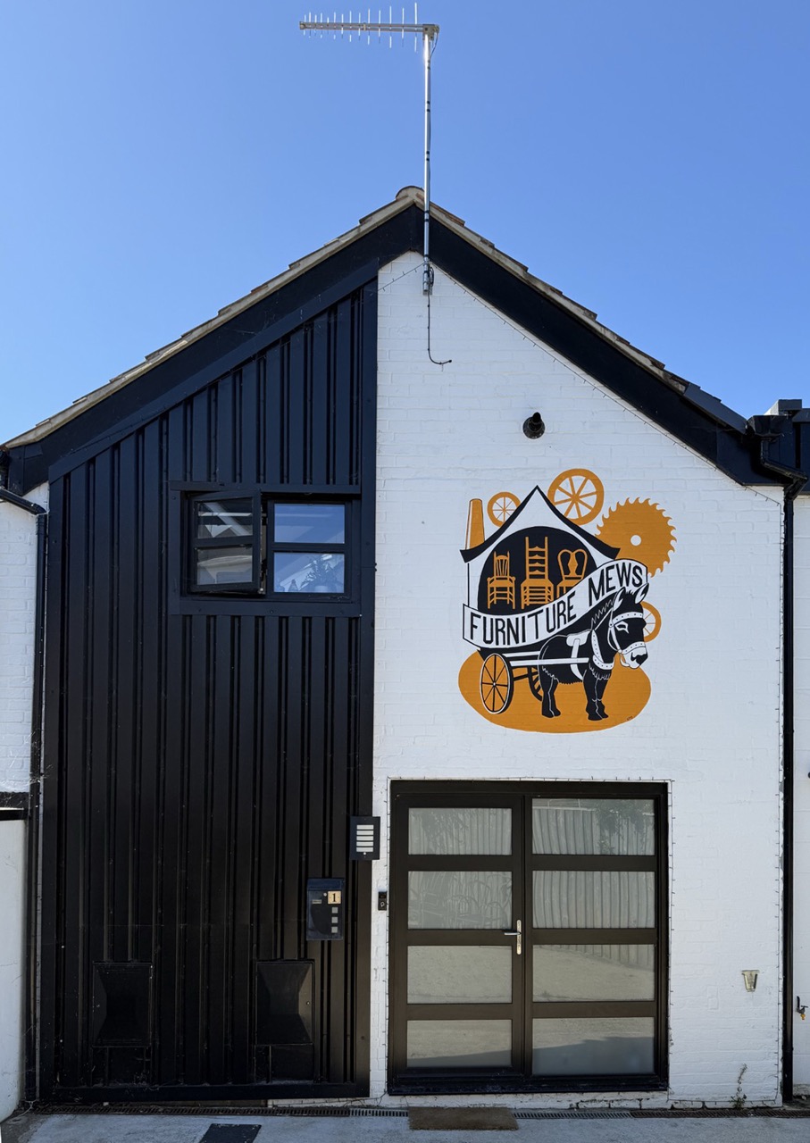 A black-and-white building facade with a large Furniture News sign, featuring a horse, wheels and gears, beneath a pitched roof and TV aerial.