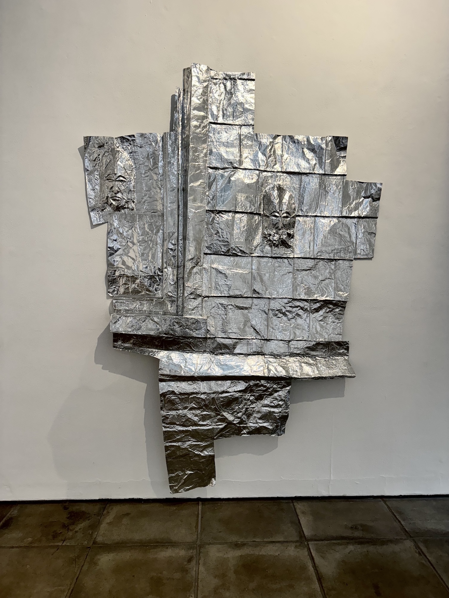 Wall-mounted sculpture of stacked metallic silver rectangular blocks with textured surfaces, arranged in irregular geometric formation