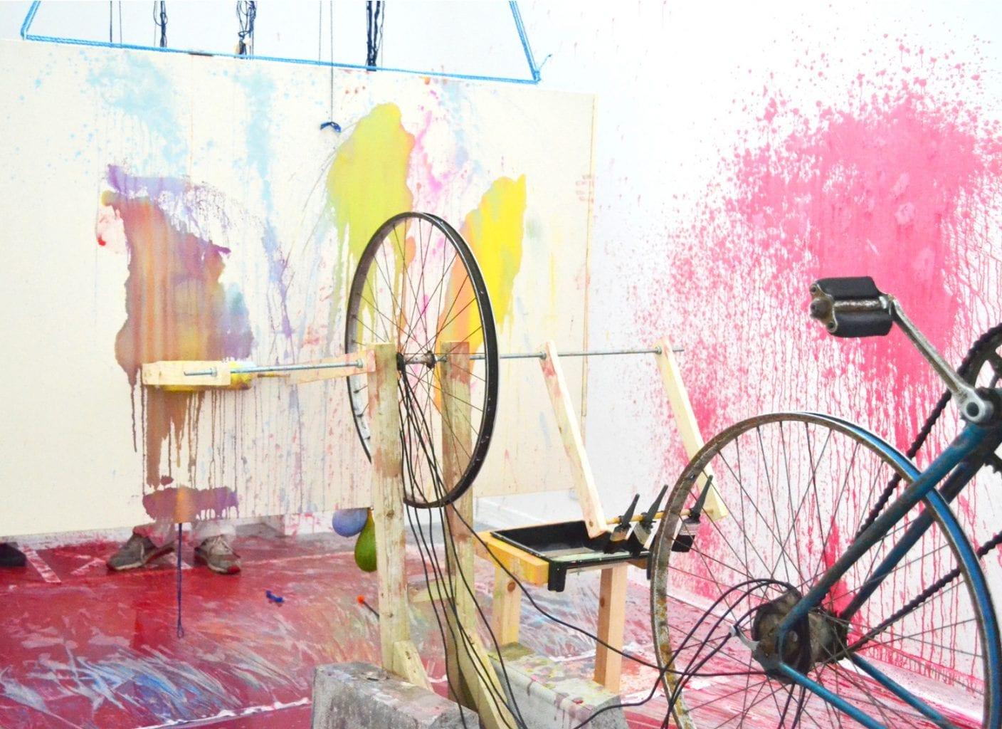 Wooden sculpture with two bicycle wheels fixed to it, sitting in a room with paint spattered on the walls.