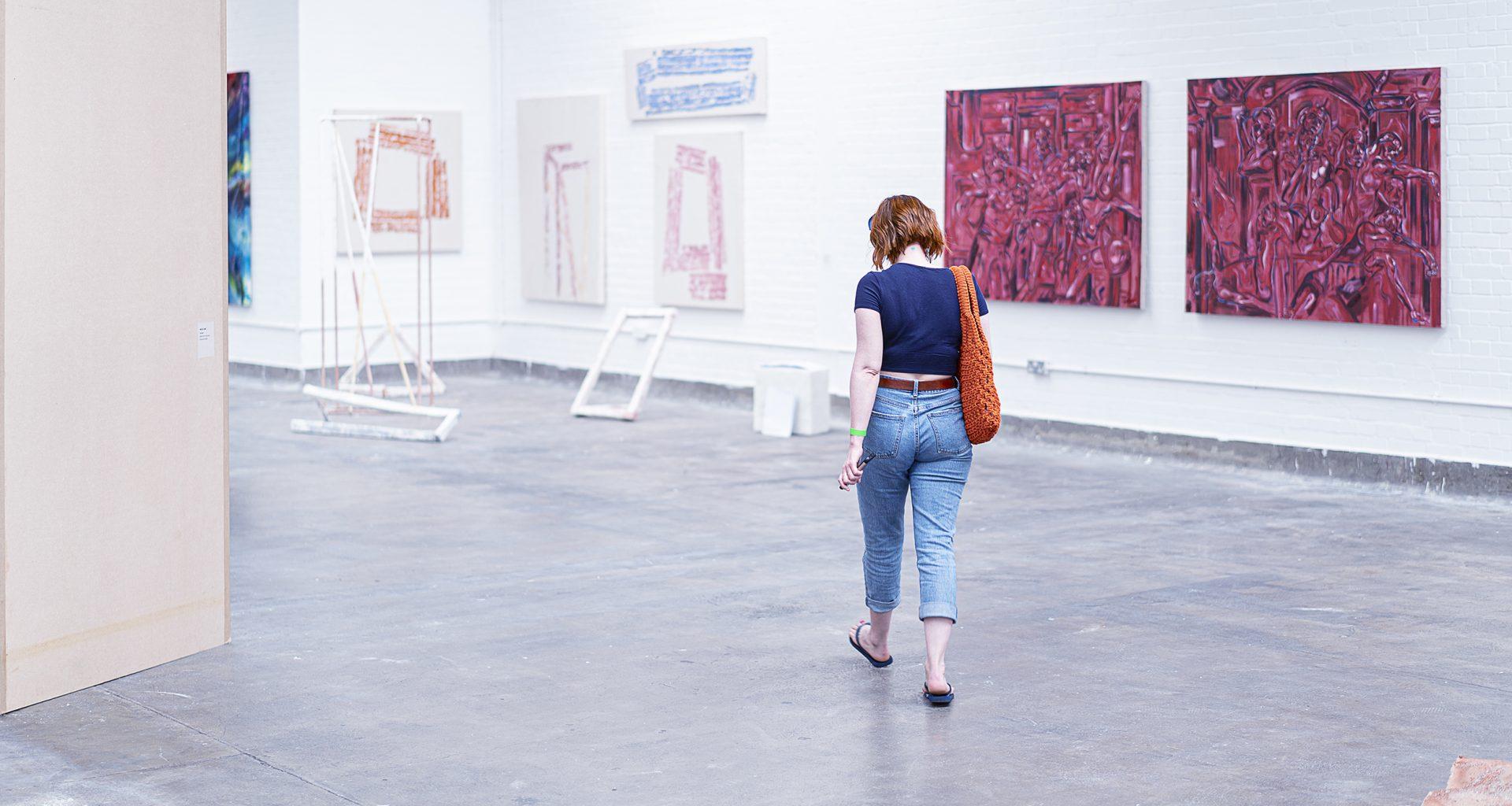 A person with their back to the camera walks through a gallery space. There are a few exhibits hanging on the wall and on stands in front of the person.
