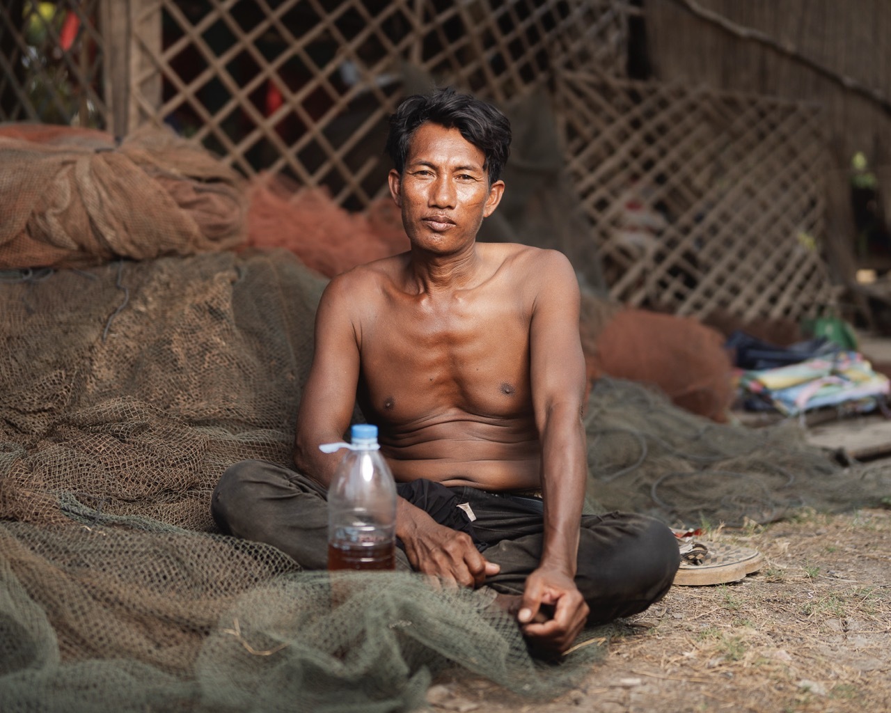 Shirtless man sits cross-legged on ground with water bottle, surrounded by sacks and lattice fencing in background.