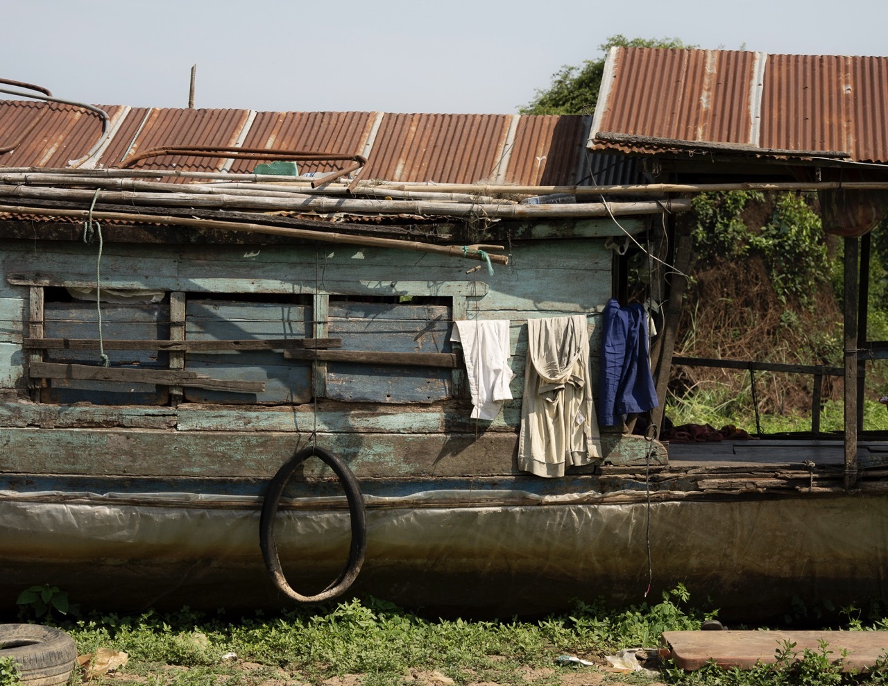 Weathered houseboat with corrugated metal roof, washing line with clothes, wooden railings, and tyre fender moored at waterside.