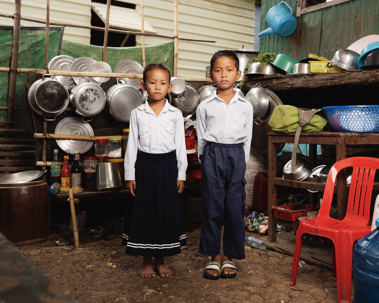 Two children in white shirts and dark trousers standing in kitchen area with metal pots, pans, and colourful plastic containers on shelves.