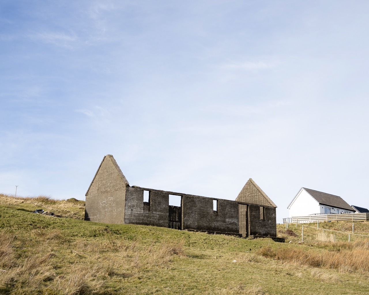 Abandoned roofless house with triangular timber gables and dark horizontal sections set on grassy hillside under blue sky with white clouds.
