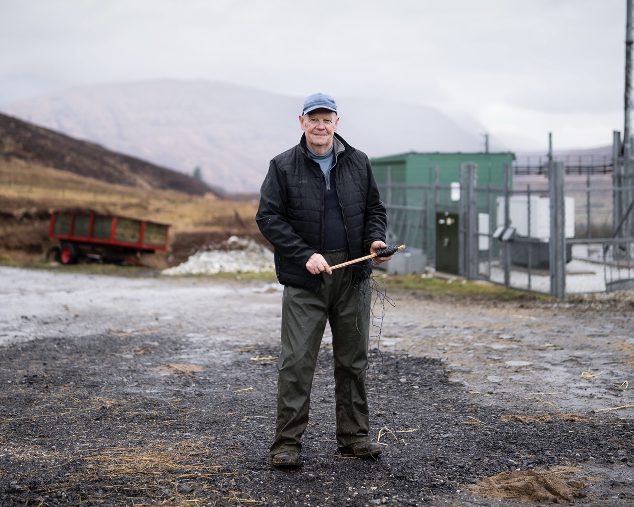 Man in dark jacket and cap holding walking stick stands on gravel ground with green industrial building and hills behind.