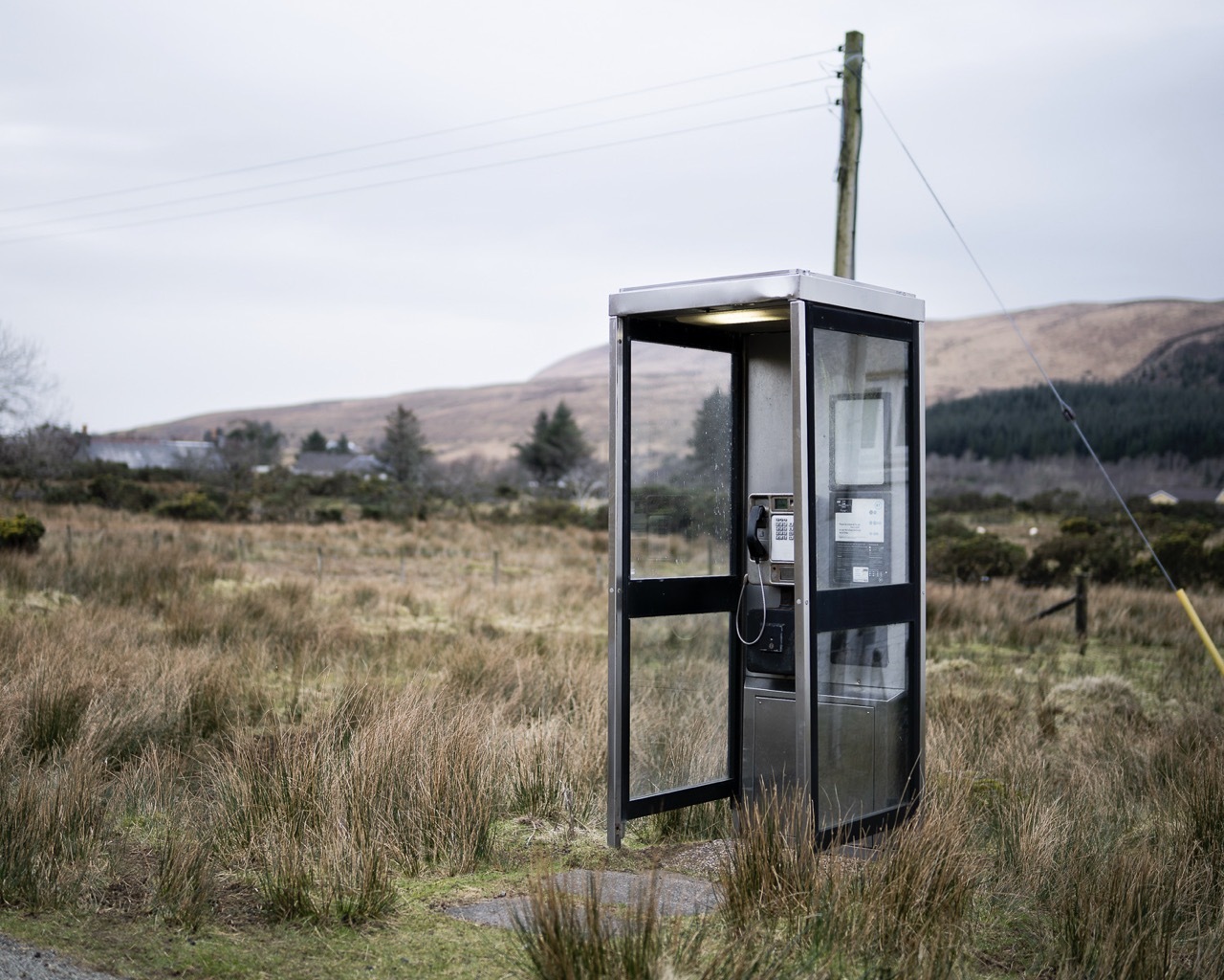 Glass telephone box in grassy field with hills and overcast sky in background, telephone pole visible behind box.