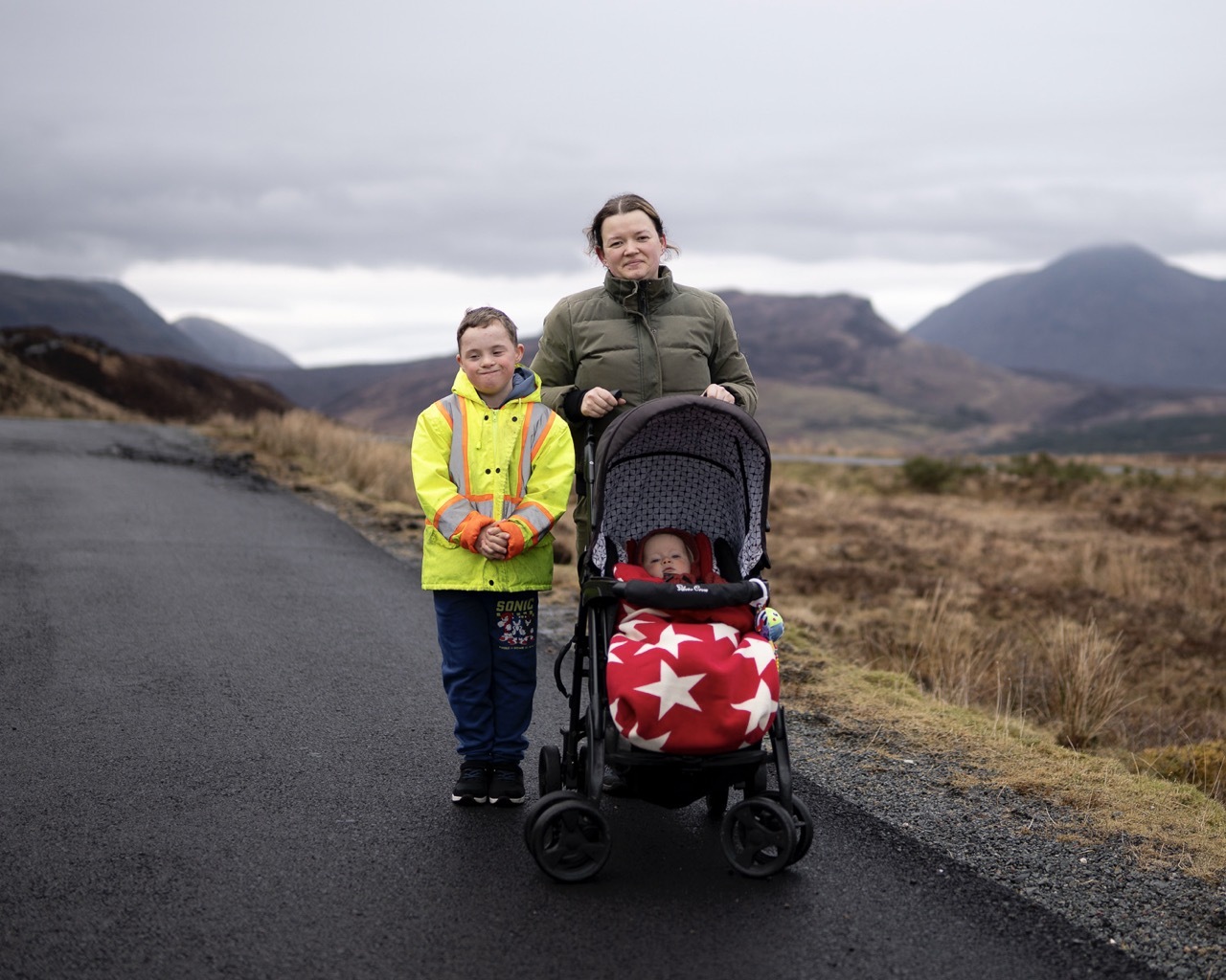 Woman in green jacket with pushchair and child in high-vis yellow jacket standing on rural road with hills in background.