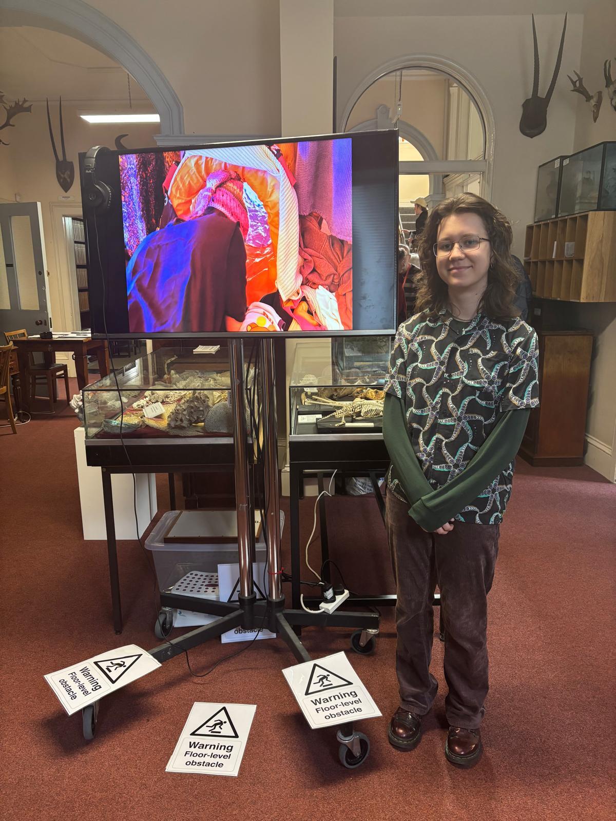 Woman standing next to television on wheeled stand in museum gallery. Screen displays colourful cave imagery. Warning signs on floor.