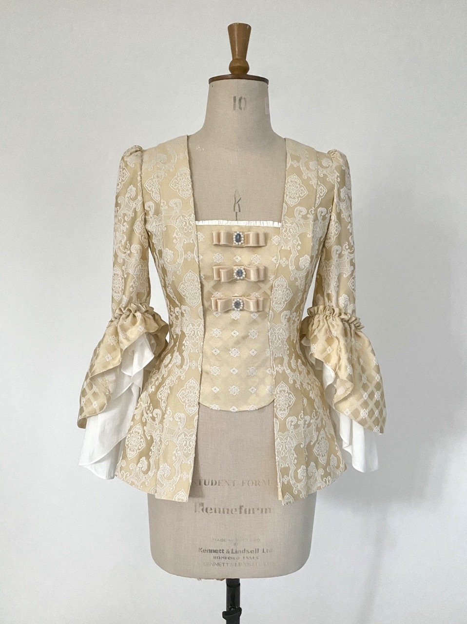 Cream-coloured period pet-en-l'air jacket with white floral lace pattern, three bow details with blue gems, and puffy sleeves on mannequin.