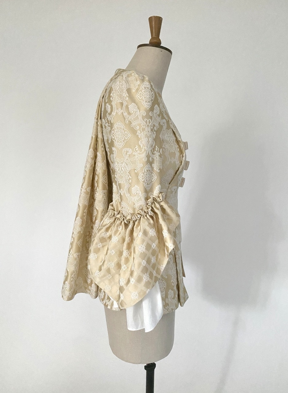 Cream-coloured period pet-en-l'air jacket with white floral lace pattern, three bow details with blue gems, and puffy sleeves on mannequin, right side view.