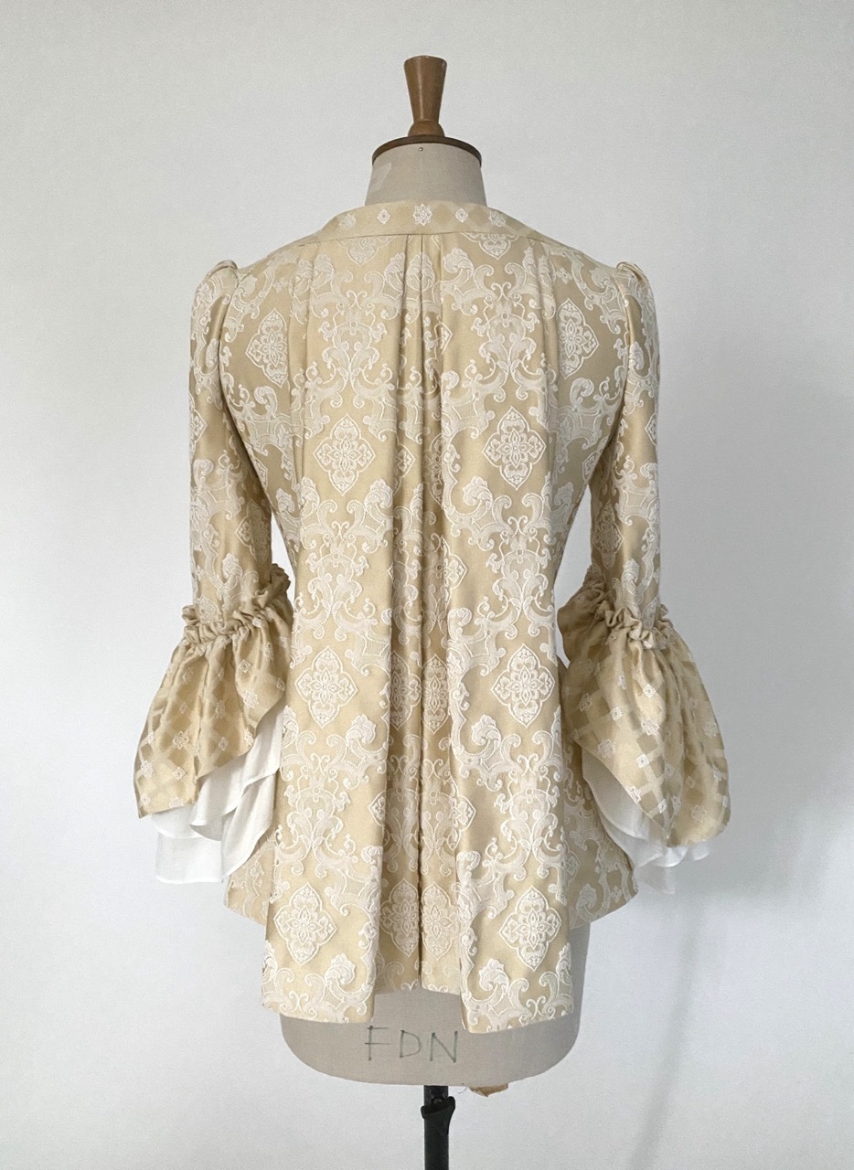 Cream-coloured period pet-en-l'air jacket with white floral lace pattern, three bow details with blue gems, and puffy sleeves on mannequin, back view.