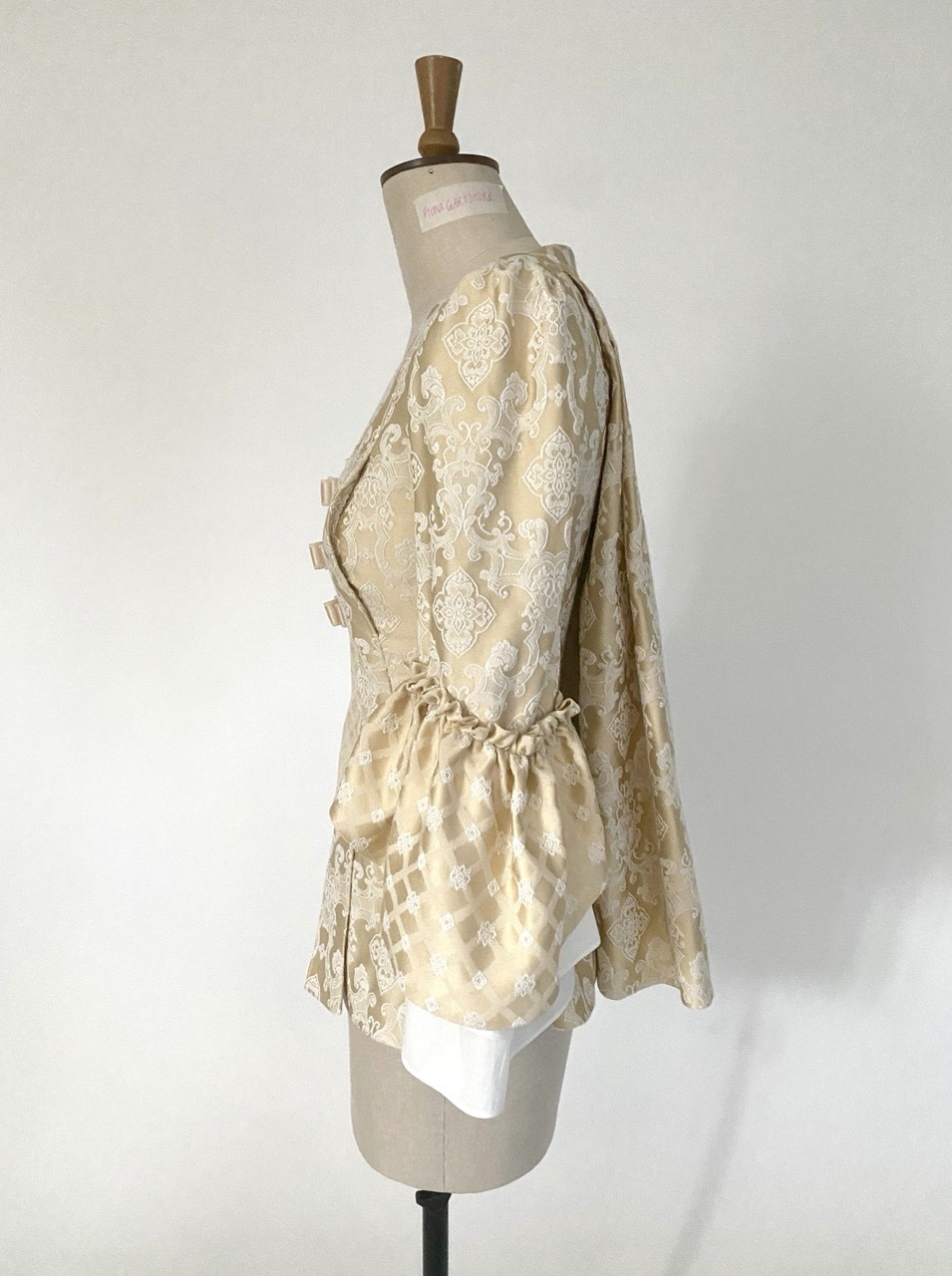 Cream-coloured period pet-en-l'air jacket with white floral lace pattern, three bow details with blue gems, and puffy sleeves on mannequin, left side view.