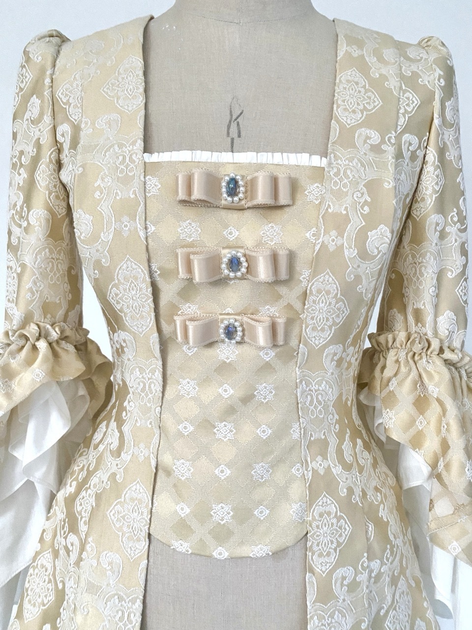 Cream-coloured period pet-en-l'air jacket with white floral lace pattern, three bow details with blue gems, and puffy sleeves on mannequin.