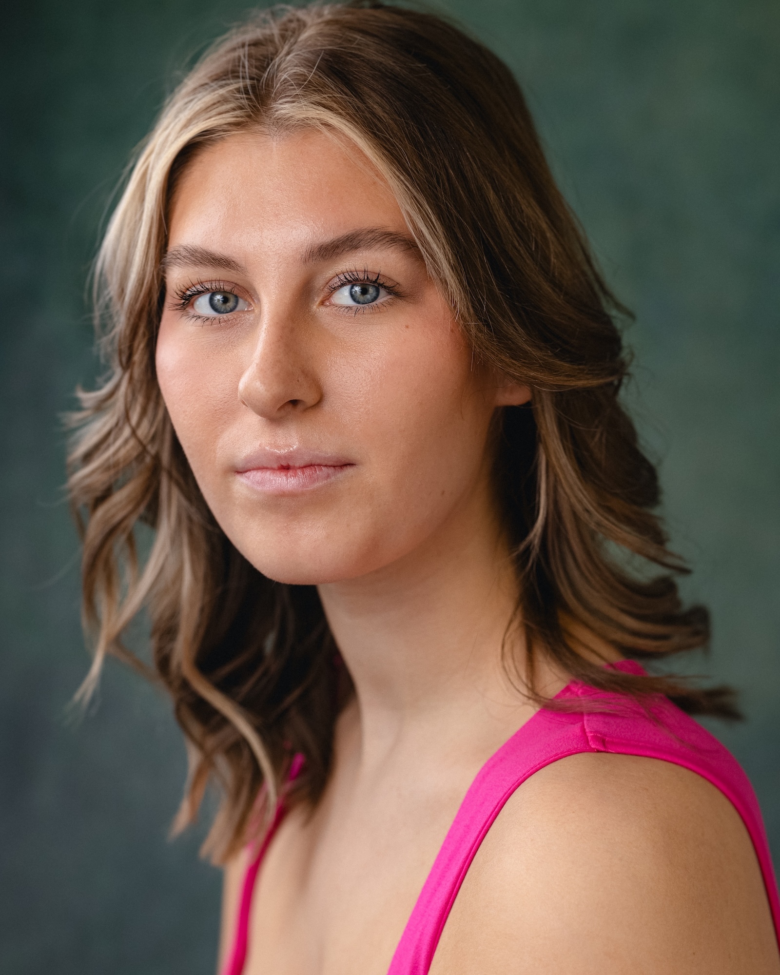 Woman with shoulder-length blonde wavy hair and blue eyes wearing bright pink sleeveless top against blurred green background.