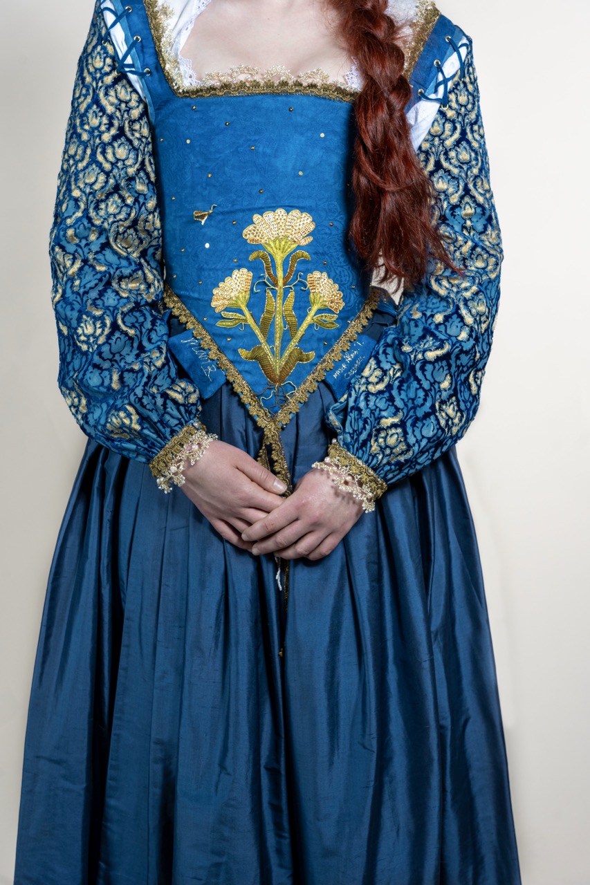 A woman wearing a blue medieval-style dress with gold embroidery and lace sleeves stands against a plain light background.