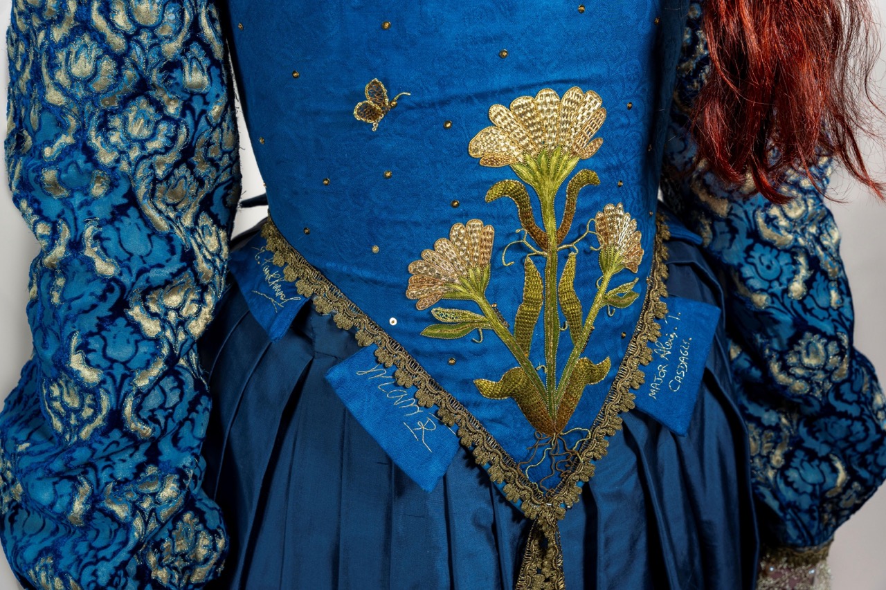 A blue embroidered bodice with gold floral detailing and matching patterned sleeves, worn over a pleated skirt.