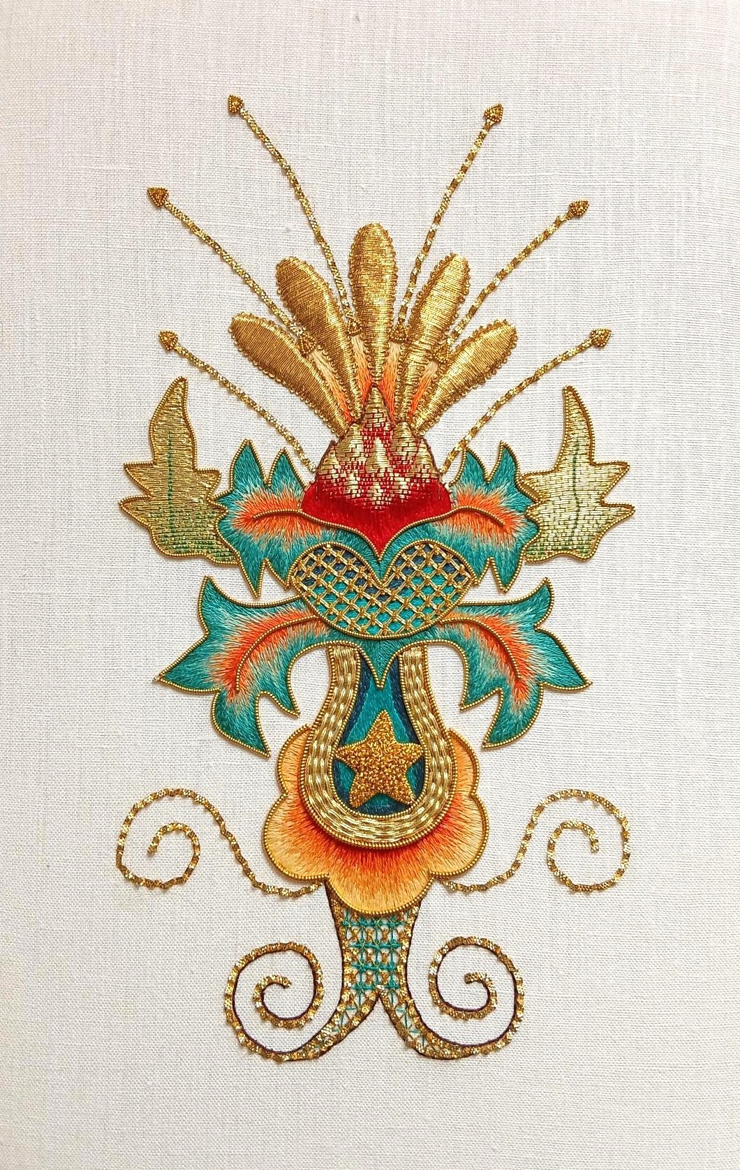 An ornate embroidered motif in gold, teal and red with leaf-like shapes and curling flourishes on a light fabric background.