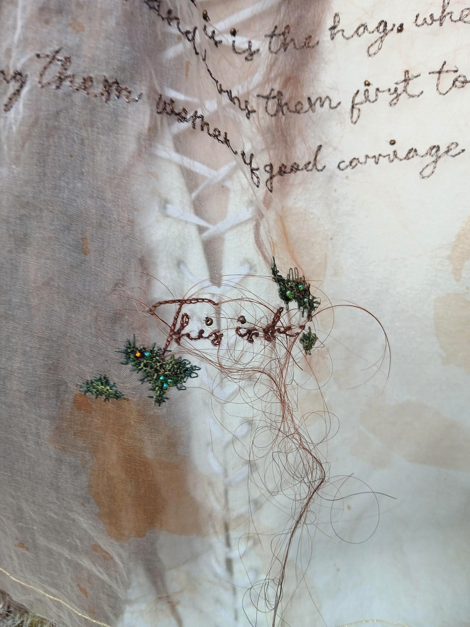 A close-up of sheer beige fabric with handwritten cursive text and stitched green floral details, with loose tan threads and abstract pale shapes in the background.