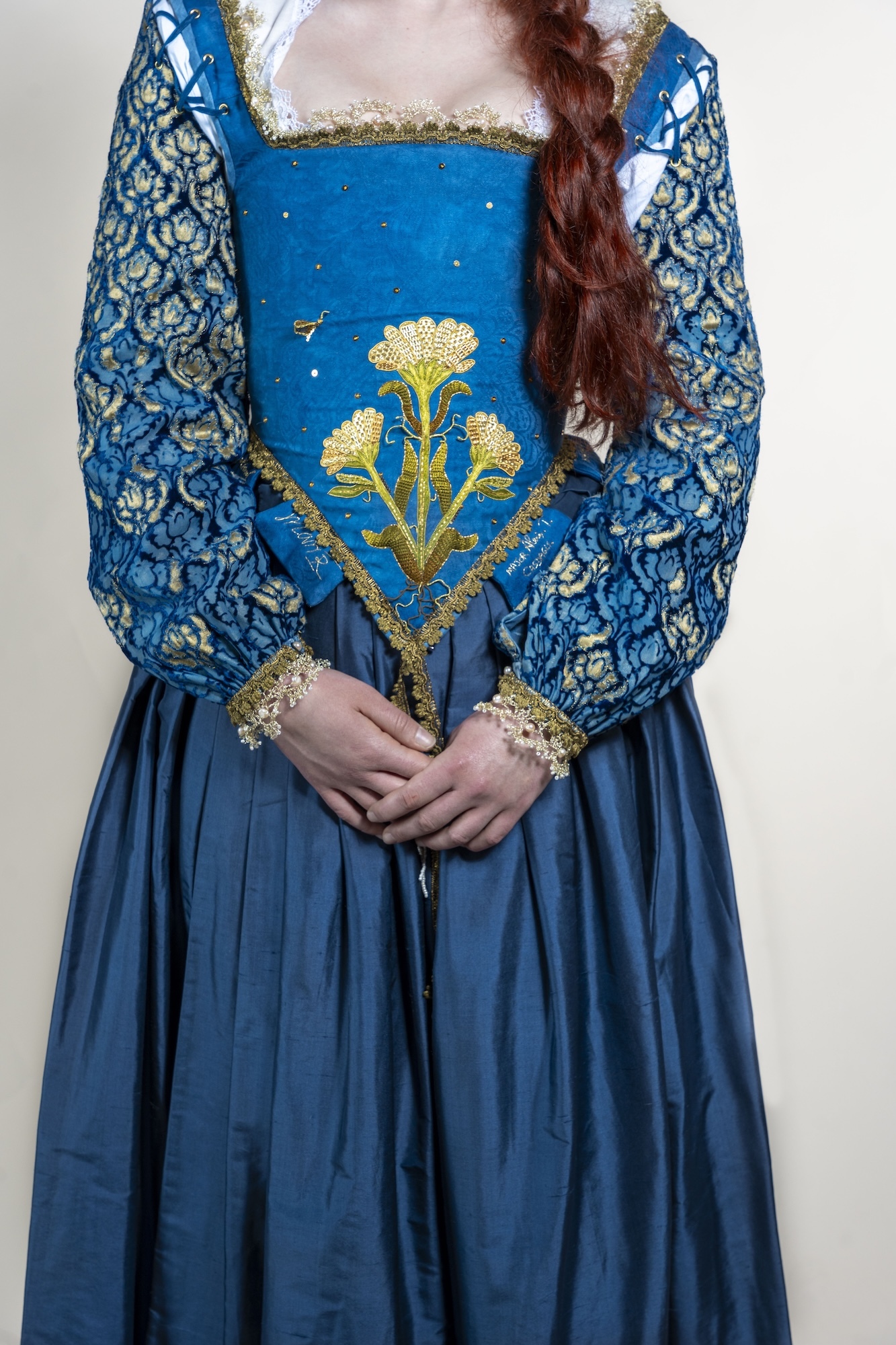 Woman in blue period dress with gold floral embroidery, patterned sleeves, white lace collar, and long auburn plait.