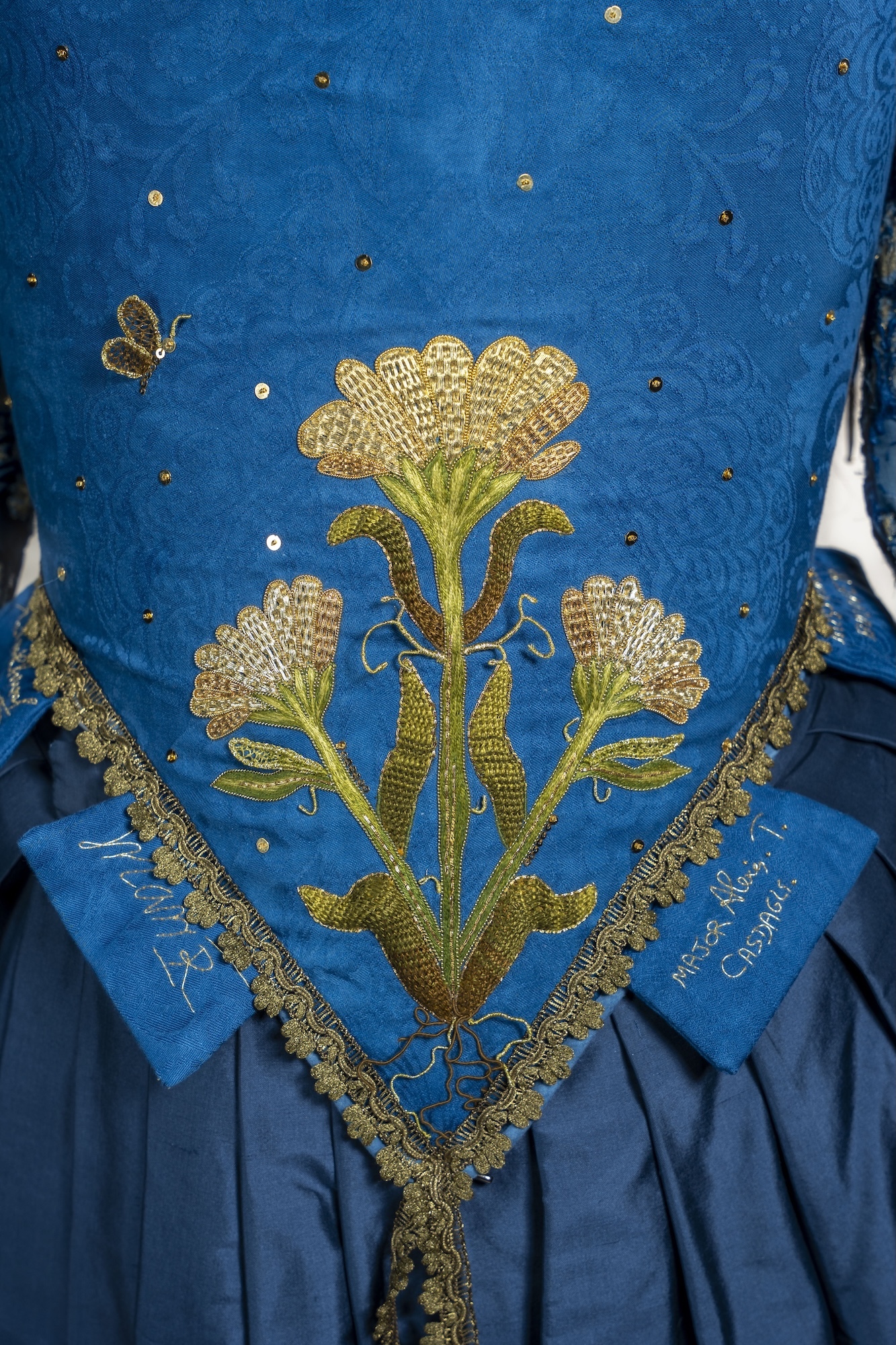 Blue silk garment with gold embroidered floral motifs, decorative trim, scattered sequins, and small bee embellishment.