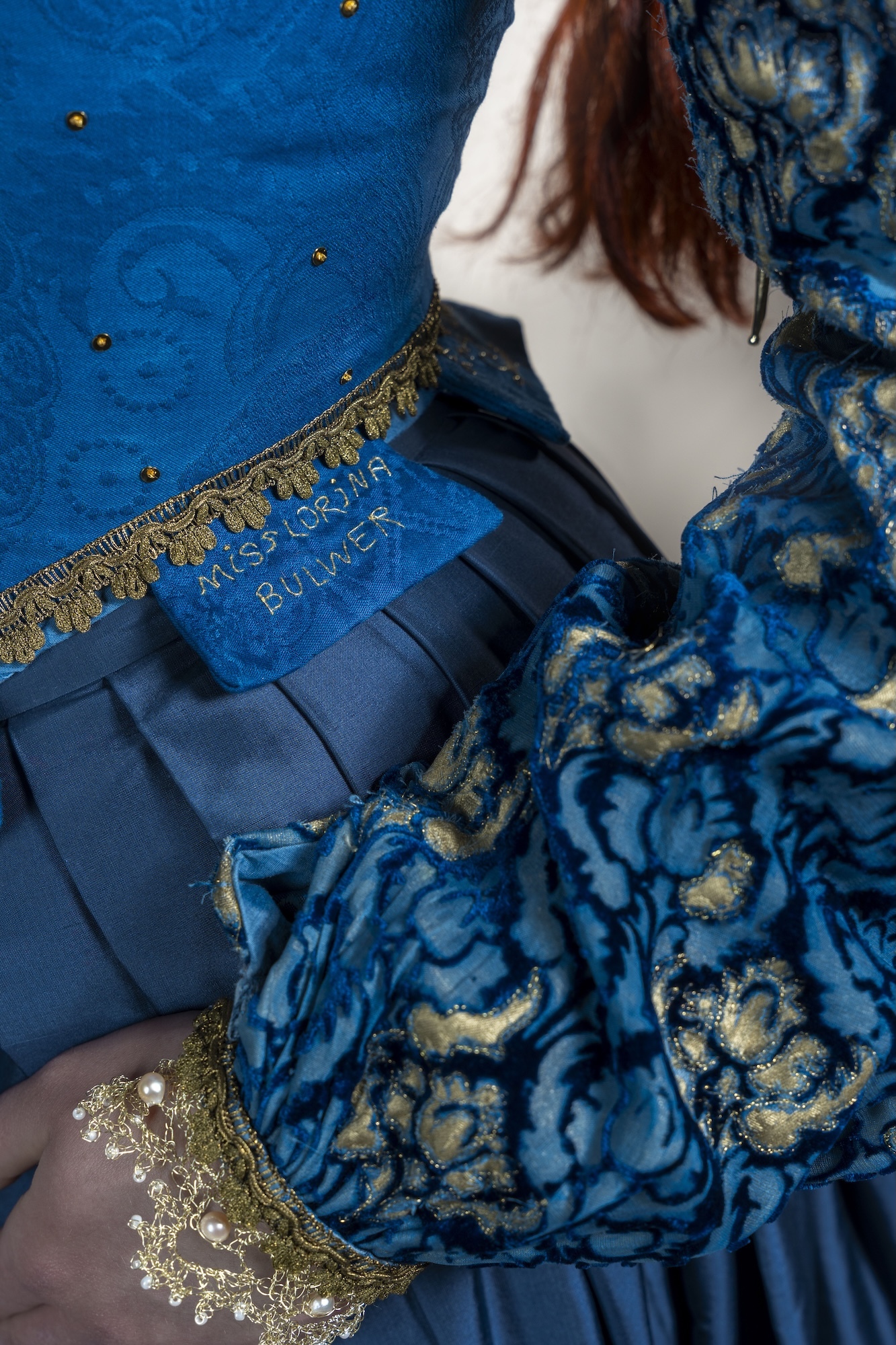Person in blue and gold floral dress with dark hair, holding blue book with gold trim against blue fabric background.