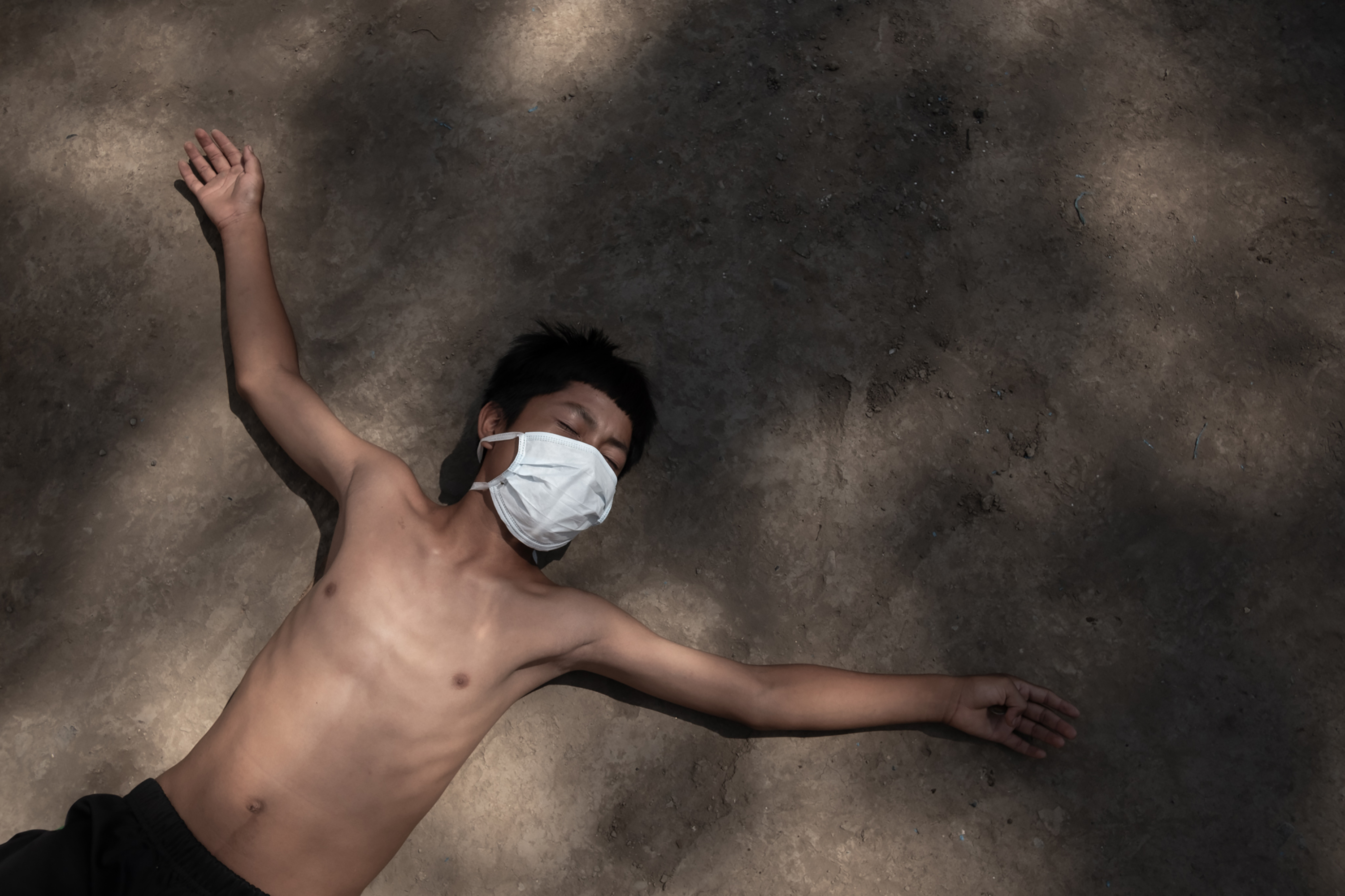 A shirtless man wearing a face mask lies on a beach in partial shade with his arms outstretched