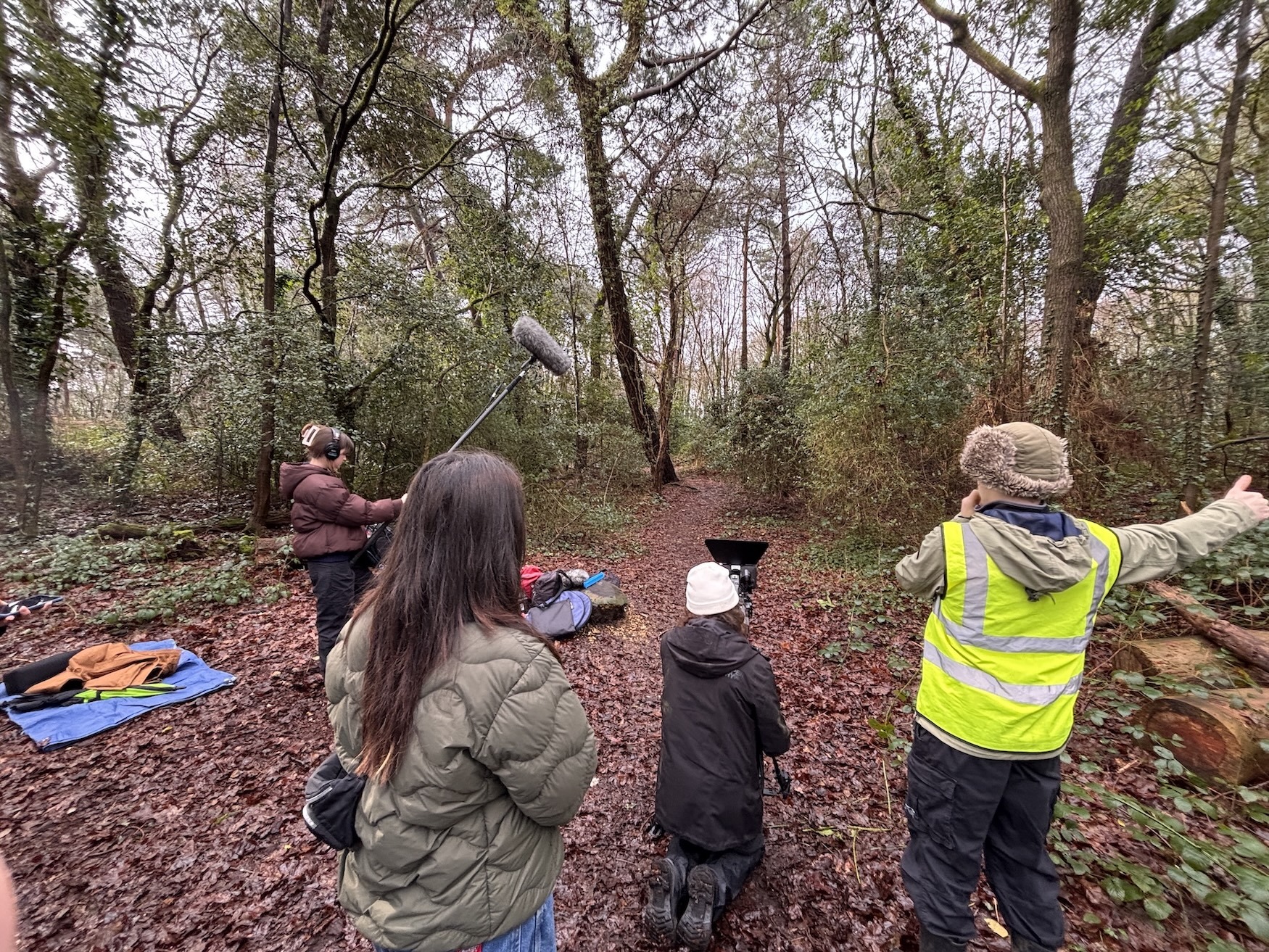 Group of people in woodland clearing with bare trees, autumn leaves on ground, person in high-vis jacket, scattered equipment visible.