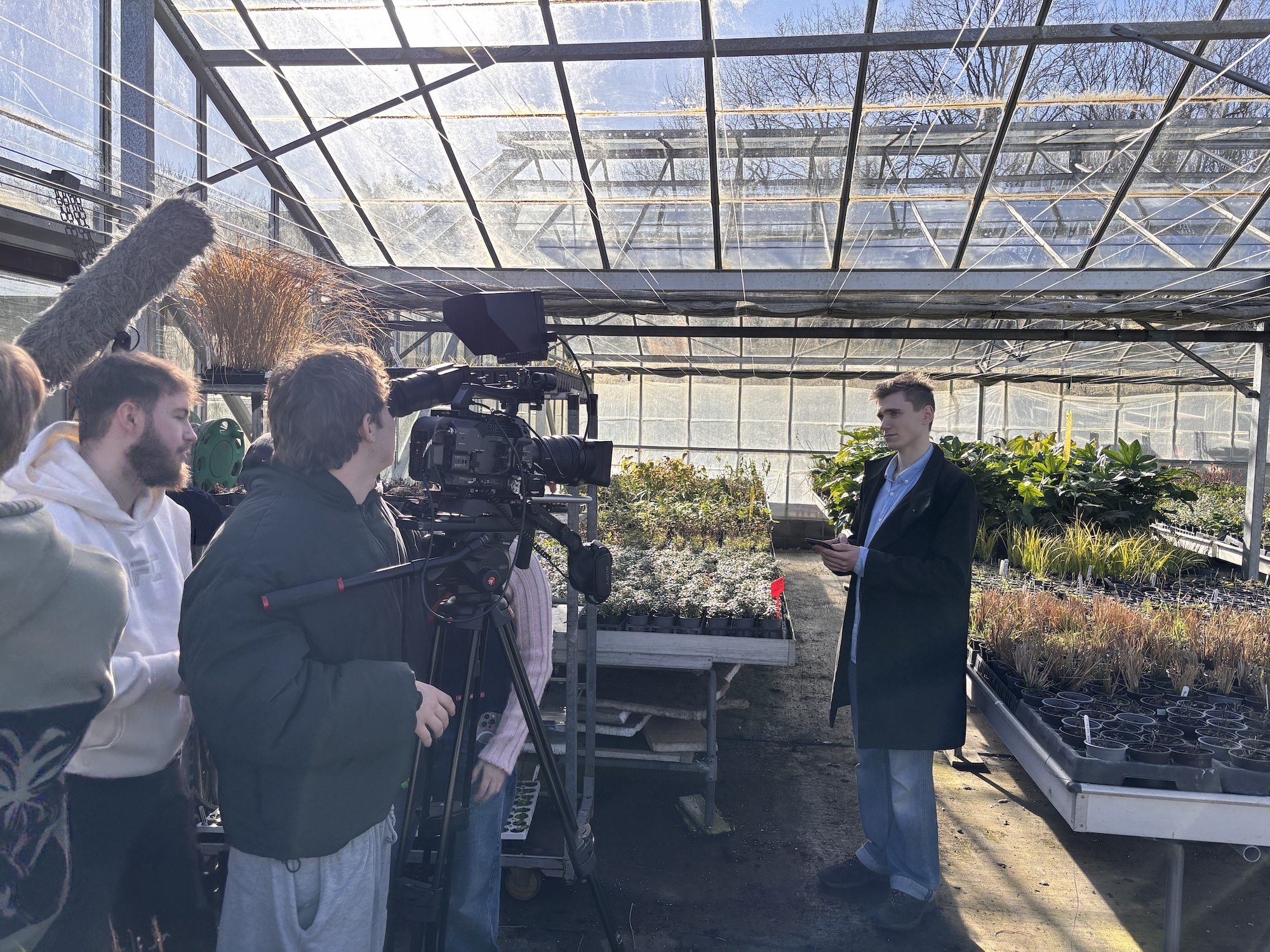Film crew with camera equipment interviewing man in suit inside glass greenhouse with plants and metal benches.