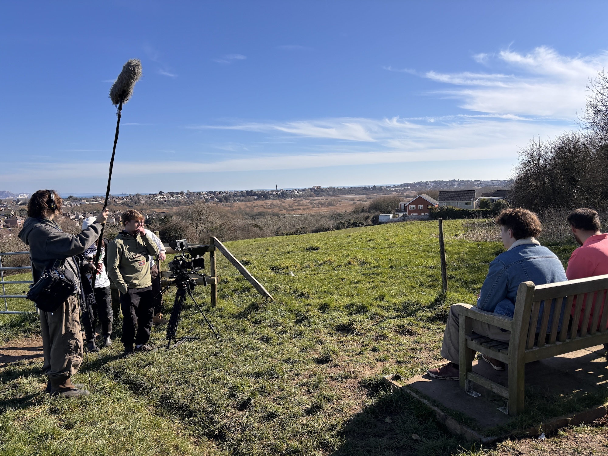 Film crew with cameras and boom microphone recording in green field, with two people seated on wooden bench, blue sky background.