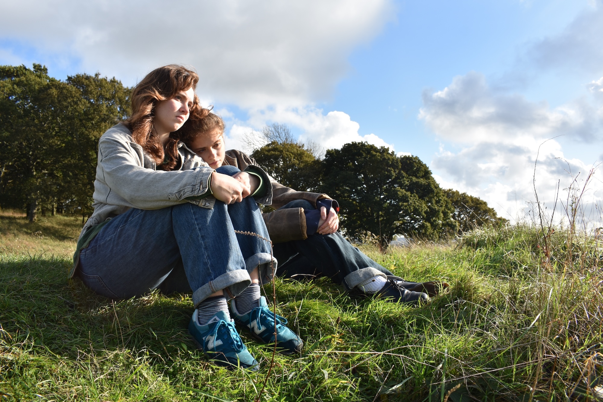 Actors M Harlan and M Marszal sitting on a hill, looking off into the distance. One has their head on the other's shoulder.