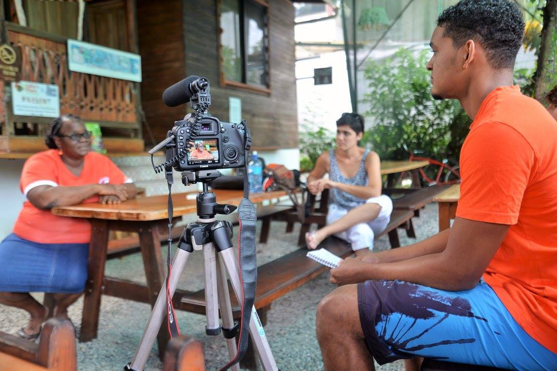 A student sitting behind a camera on a tripod while interviewing an older woman sitting on the edge of a picnic bench.