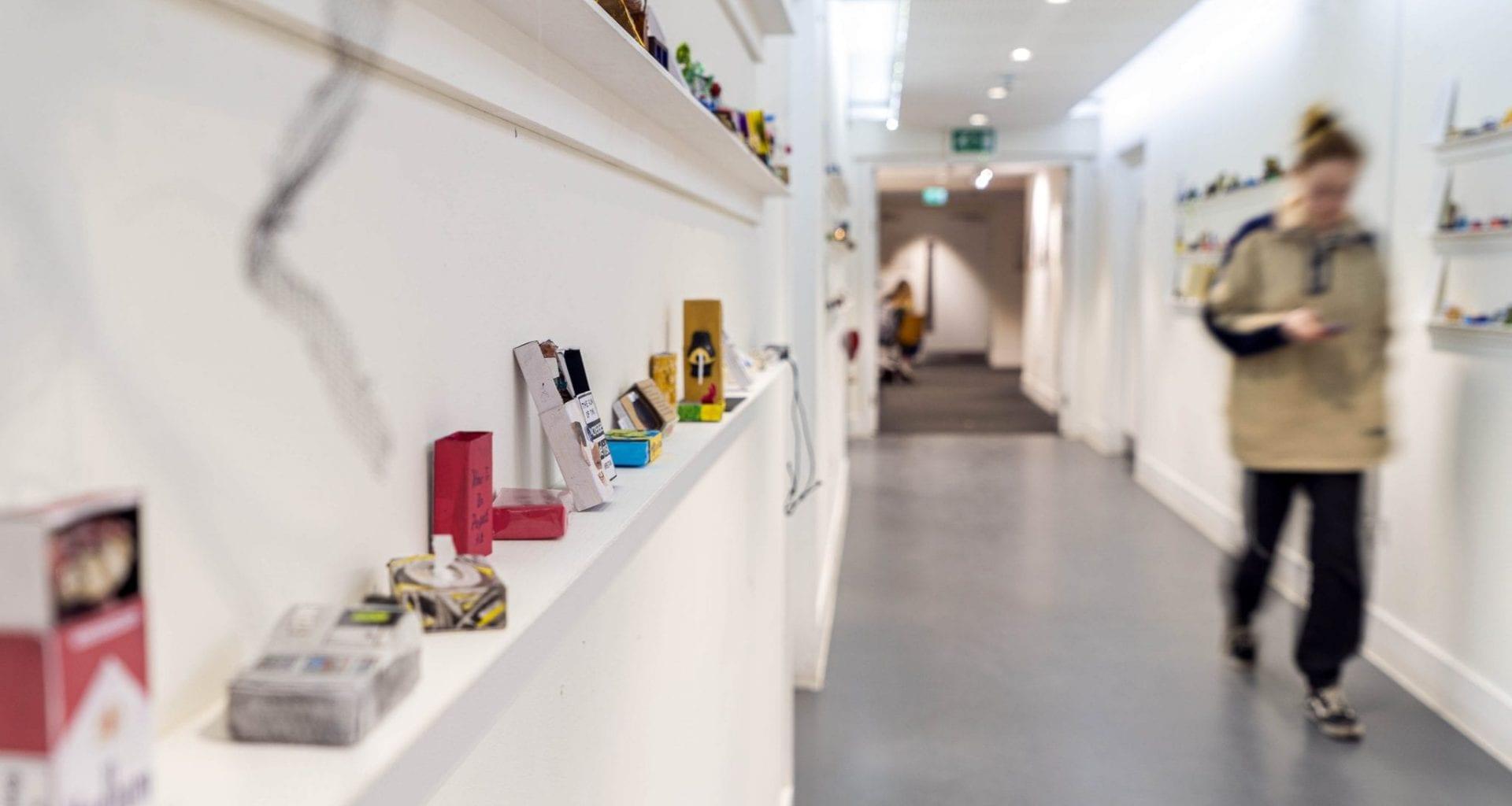 Several matchbox artworks on display in the hallway of a gallery space. A person is walking down the hallway.