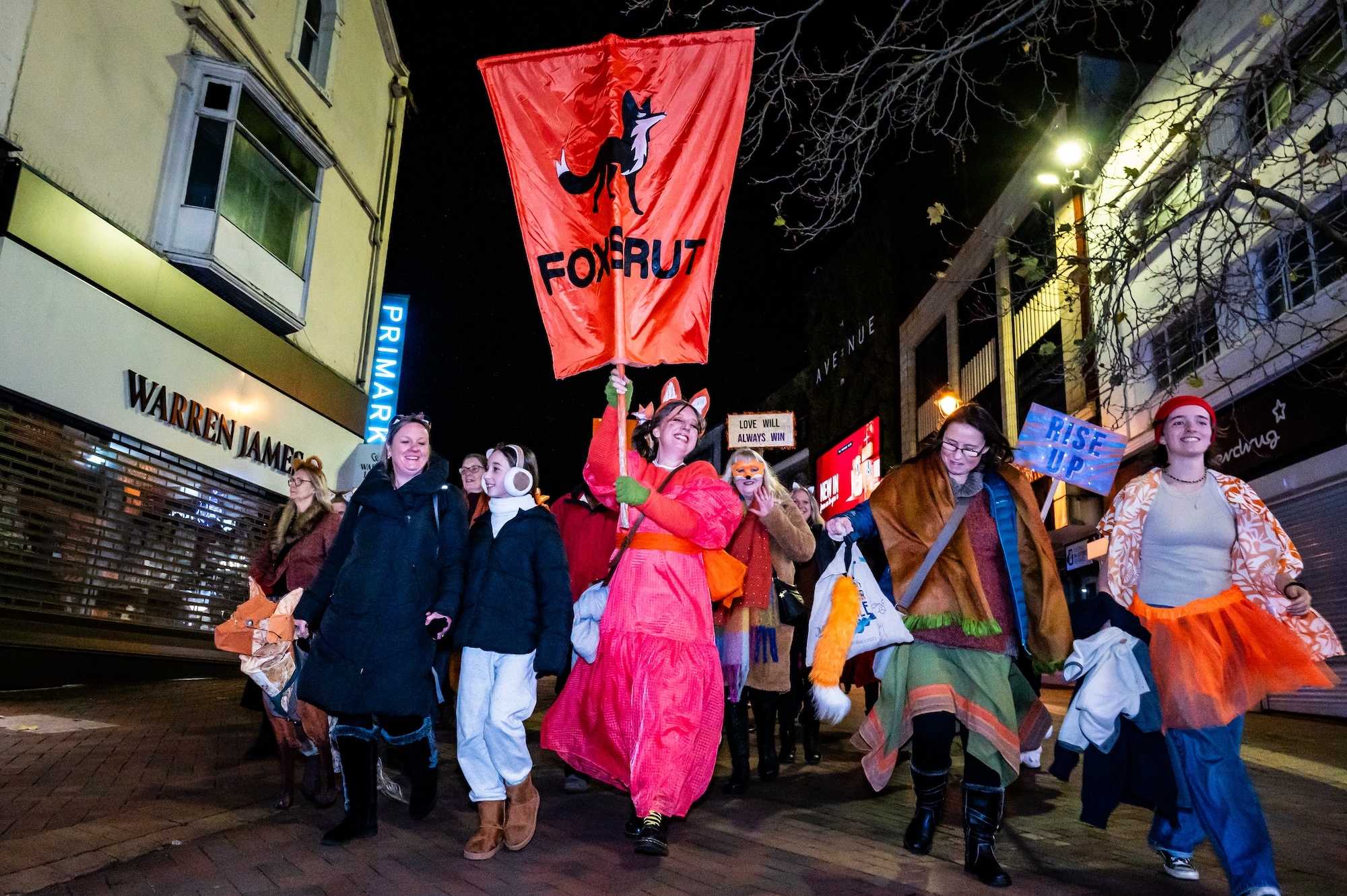 A group of people in colourful clothing walking down Bournemouth High Street. One of them is carrying a banner showing an image of a fox and the word "FOXSTRUT" underneath.