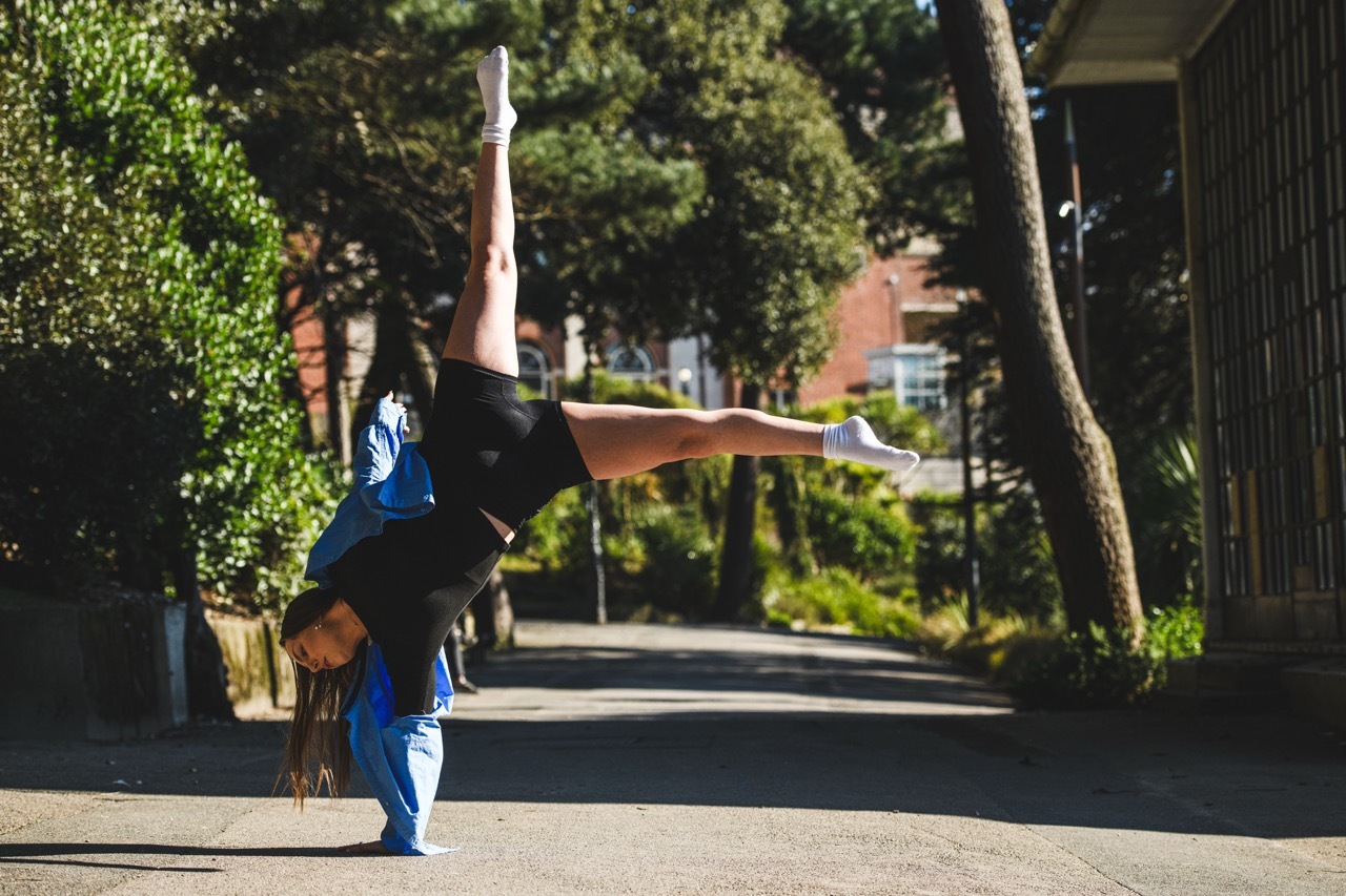 Person doing handstand on pavement with legs split apart, wearing blue top and jeans, surrounded by trees and residential buildings.