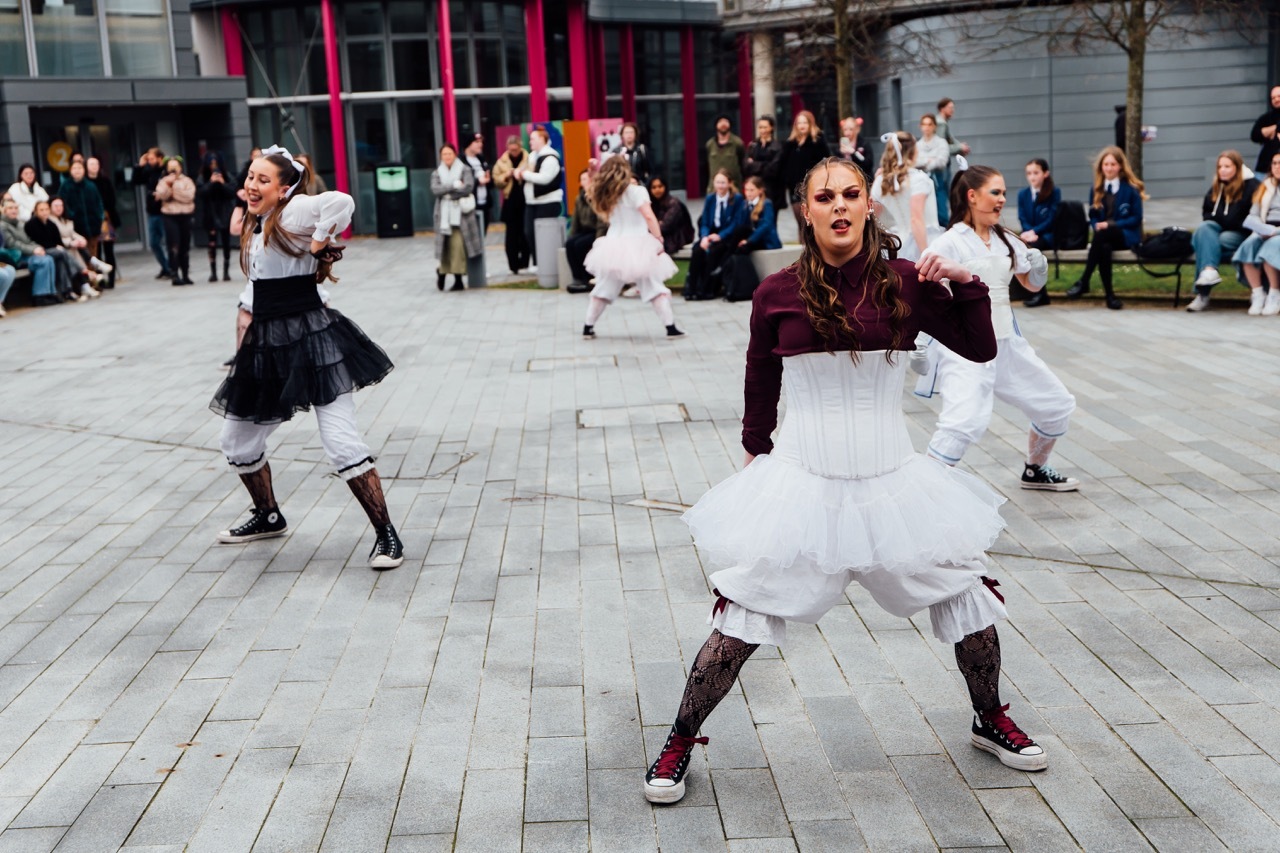 Folk dancers in traditional costumes performing on cobbled square. Women wear white skirts, dark bodices, boots. Crowd watches from modern building backdrop.